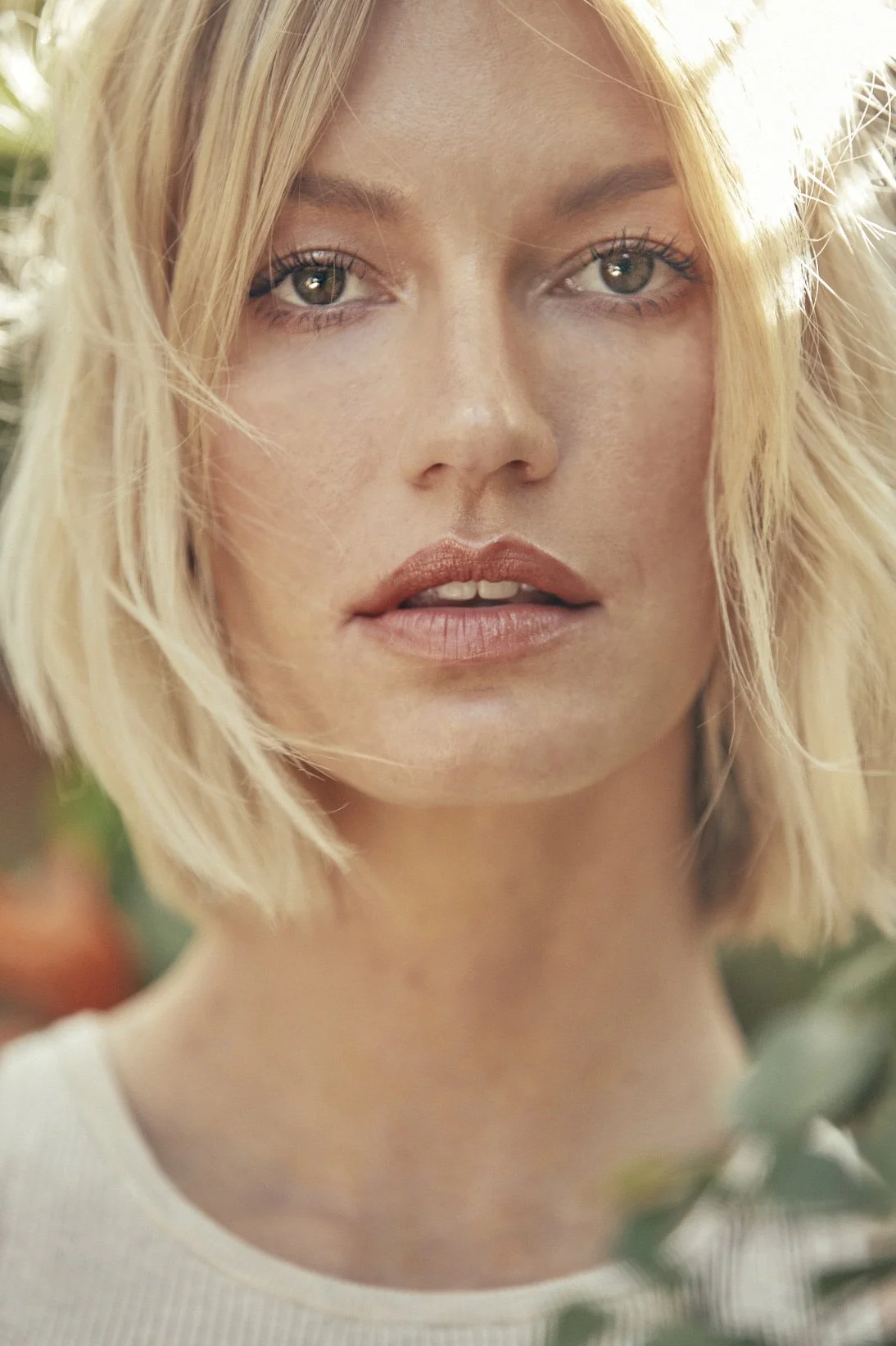 Close-up of a woman with blonde, shoulder-length hair, light makeup, and neutral expression, outdoors with blurred greenery in the background.