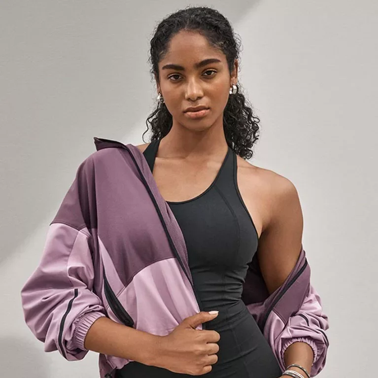 A young woman with curly dark hair wearing a black sports top and a purple jacket draped over her shoulders, standing against a plain background.