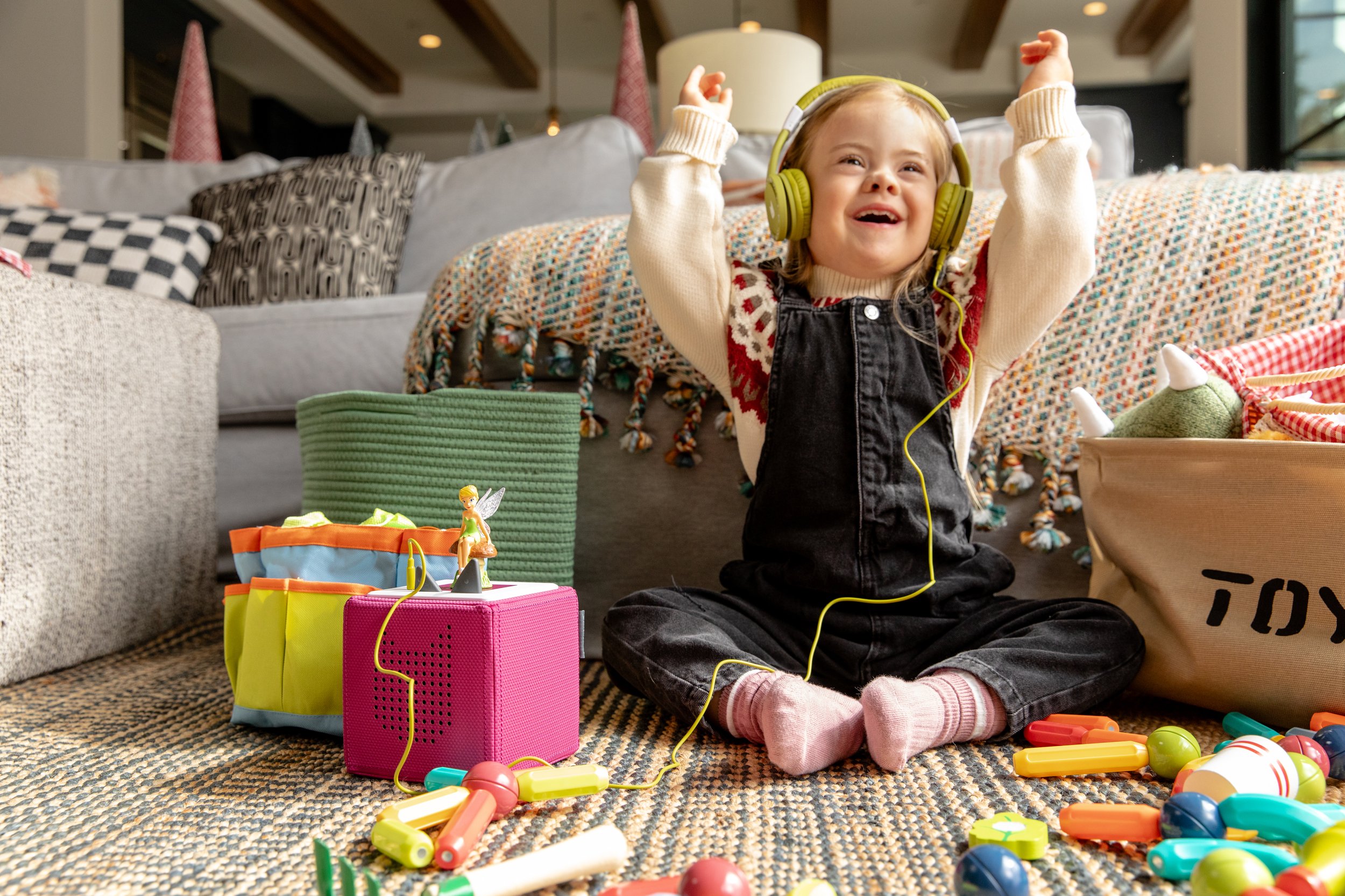A young girl sitting cross-legged on a rug, wearing headphones and smiling with her hands in the air. She is surrounded by colorful toys, including a pink speaker with a fairy figurine, a toy police light, and scattered plastic objects, in a living r