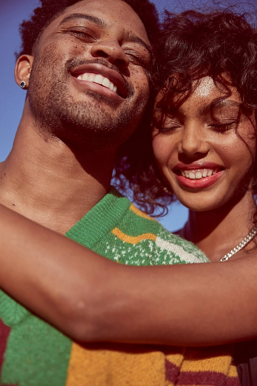A close-up of a smiling man and woman embracing, with the man showing teeth and the woman with closed eyes, both under clear blue sky.
