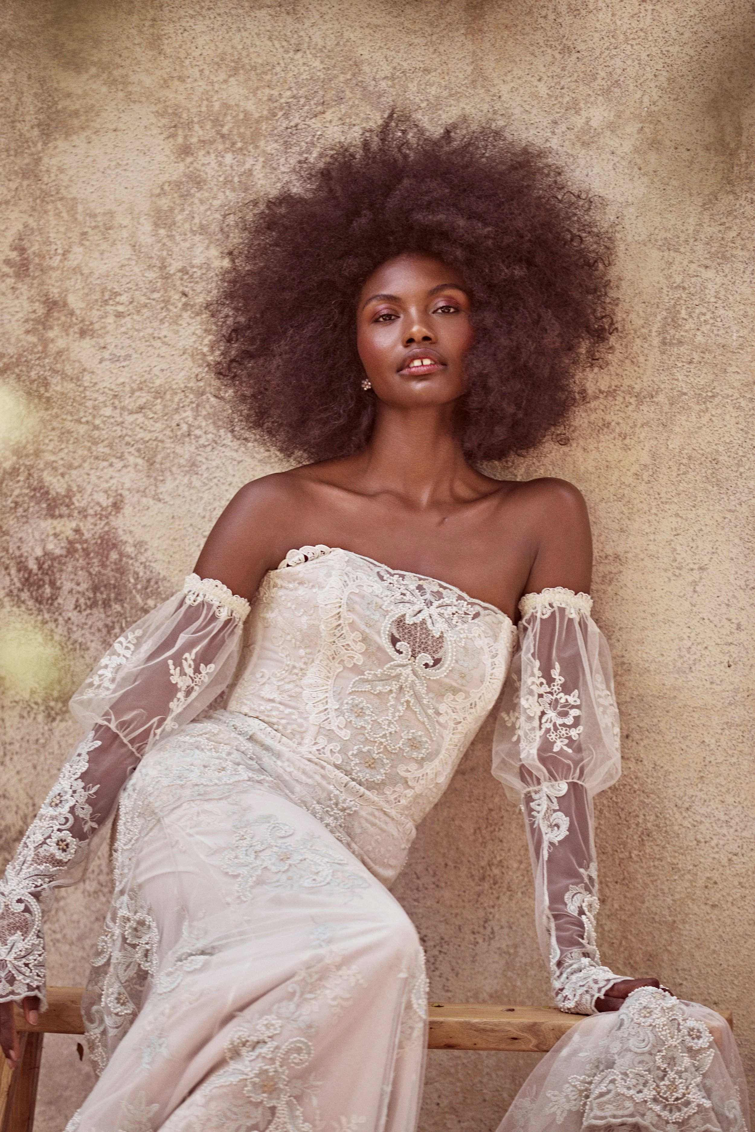 Woman with voluminous curly hair wearing an off-the-shoulder, embroidered lace dress, seated against a textured beige wall.
