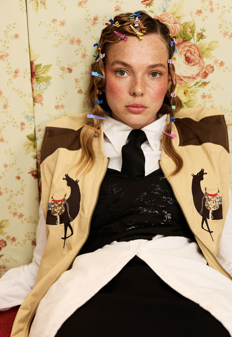 Young woman with red hair and freckles lying down, hair clipped with colorful plastic clips, wearing a cast with embroidered horses, a white shirt, black tie, and a black sequin top, against floral wallpaper.