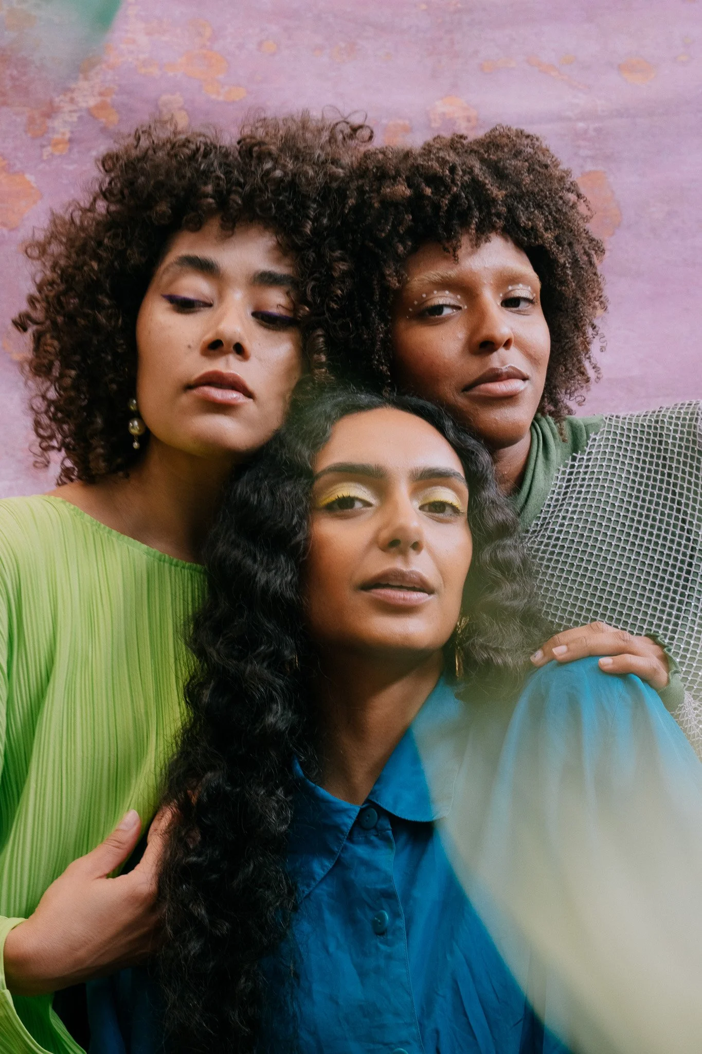 Close-up of three young women with diverse skin tones and curly hair posing together against a colorful background, wearing vibrant clothing and shiny makeup.