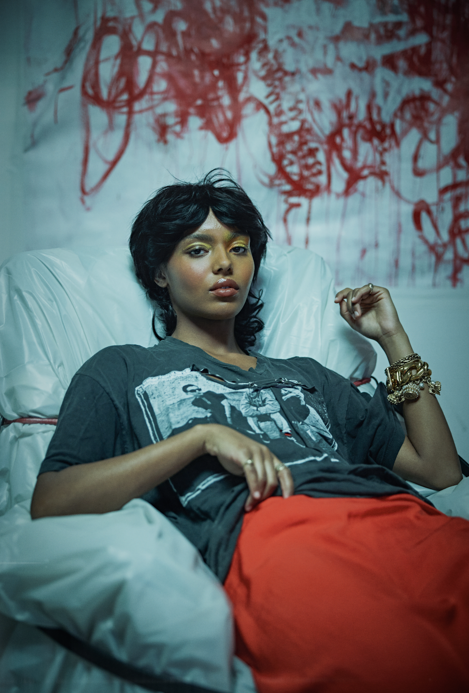 A woman with dark hair and gold eyeshadow reclining on a bed, wearing a graphic black T-shirt, red pants, and multiple gold bracelets, with a red and black abstract art piece in the background.