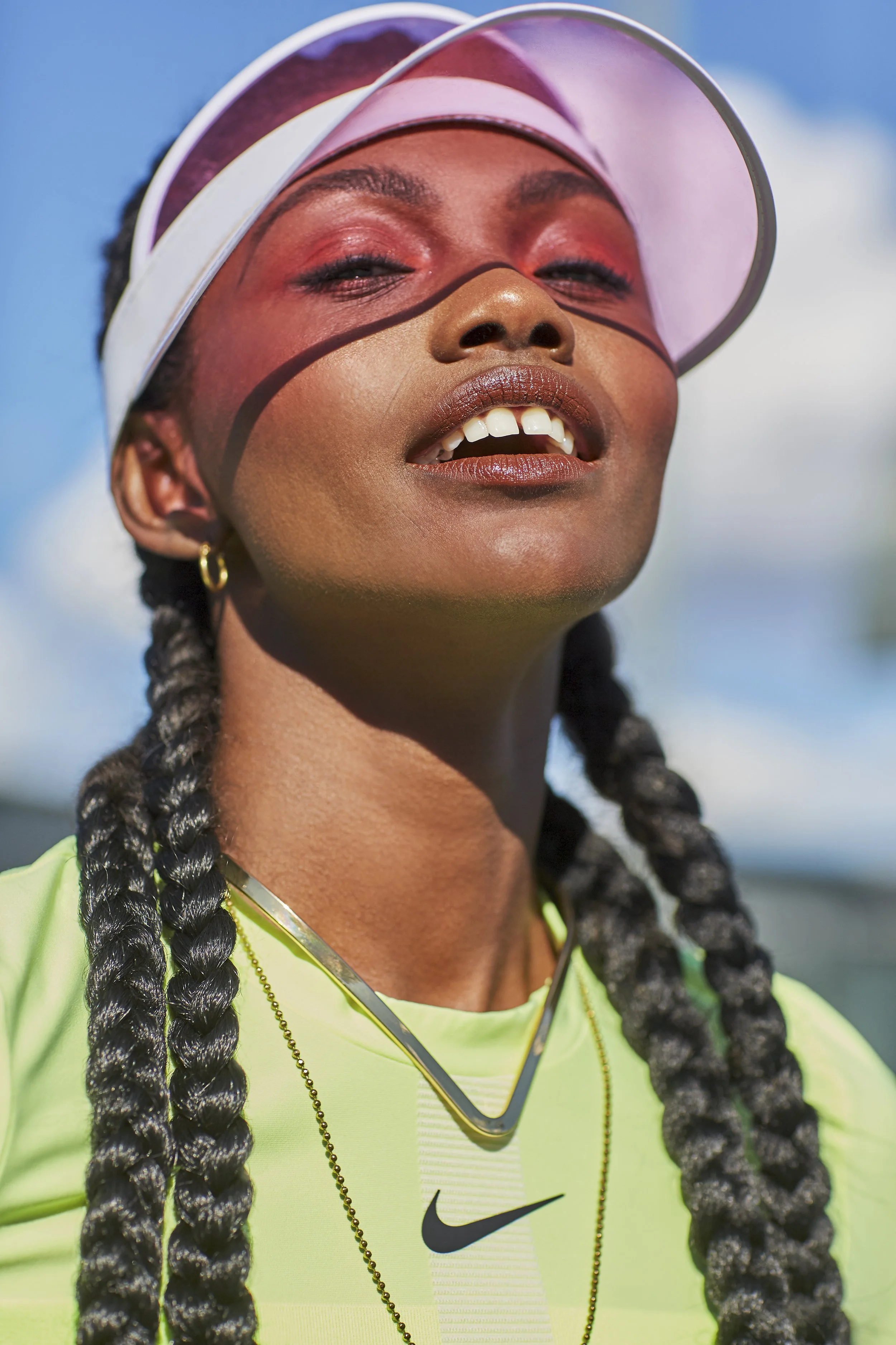 A close-up of a smiling woman with braided hair wearing a white visor, gold earrings, and a neon yellow Nike shirt outdoors against a partly cloudy sky.