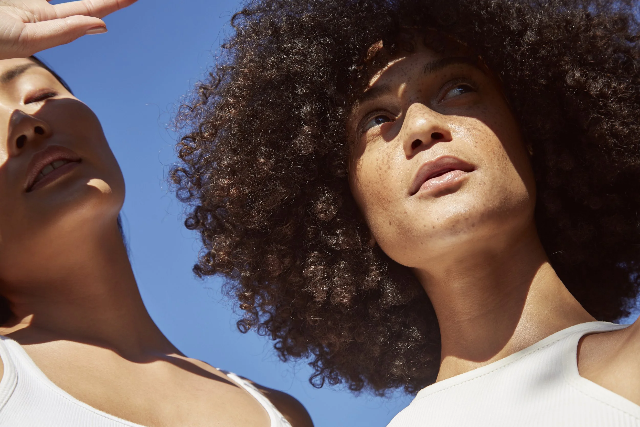 Two women with curly hair standing outdoors under a clear blue sky, looking upward and appearing to enjoy the sunlight.