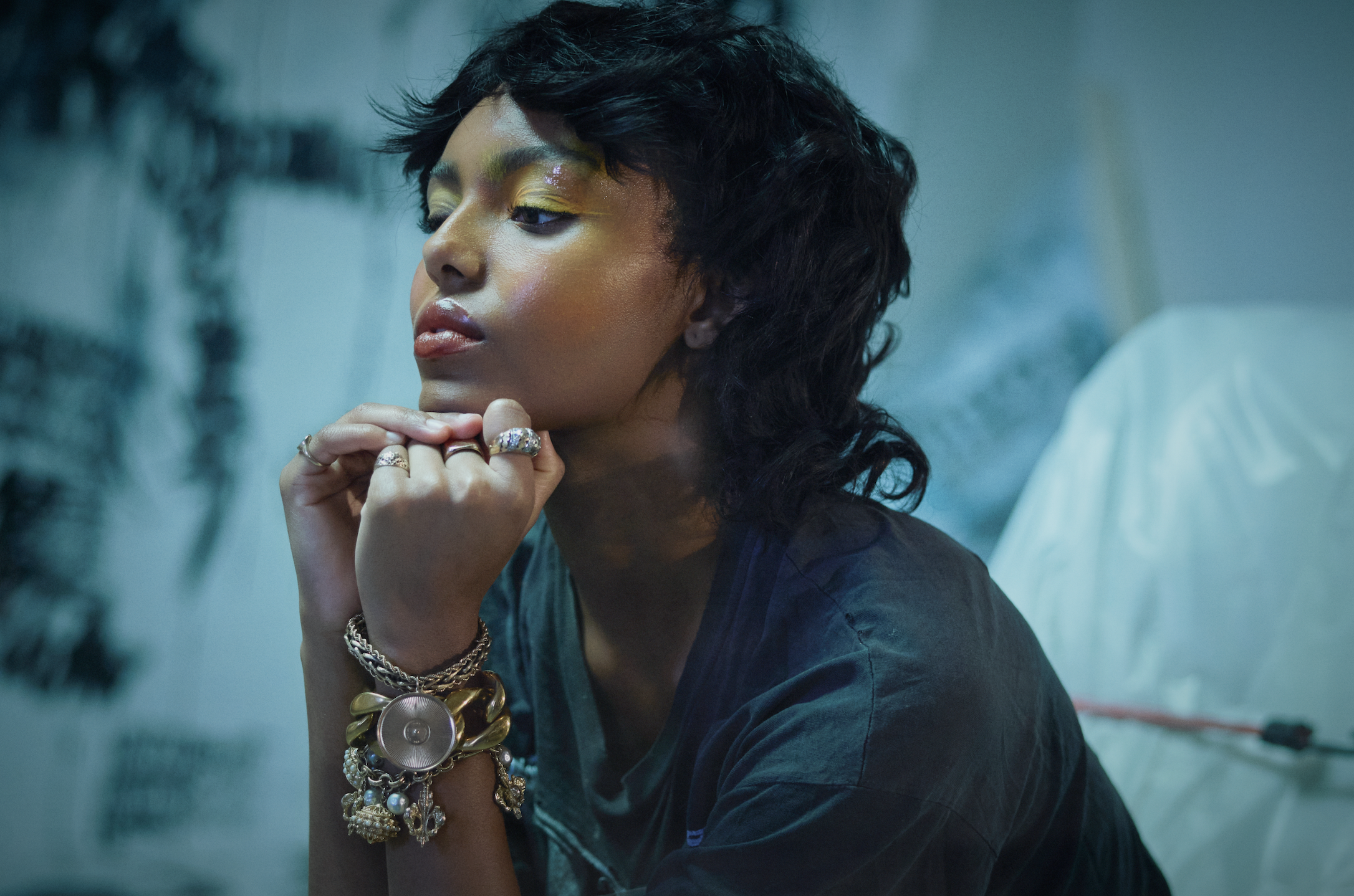 A woman with short curly black hair and colorful makeup resting her chin on her hand, wearing multiple rings and bracelets, with a blurred background.