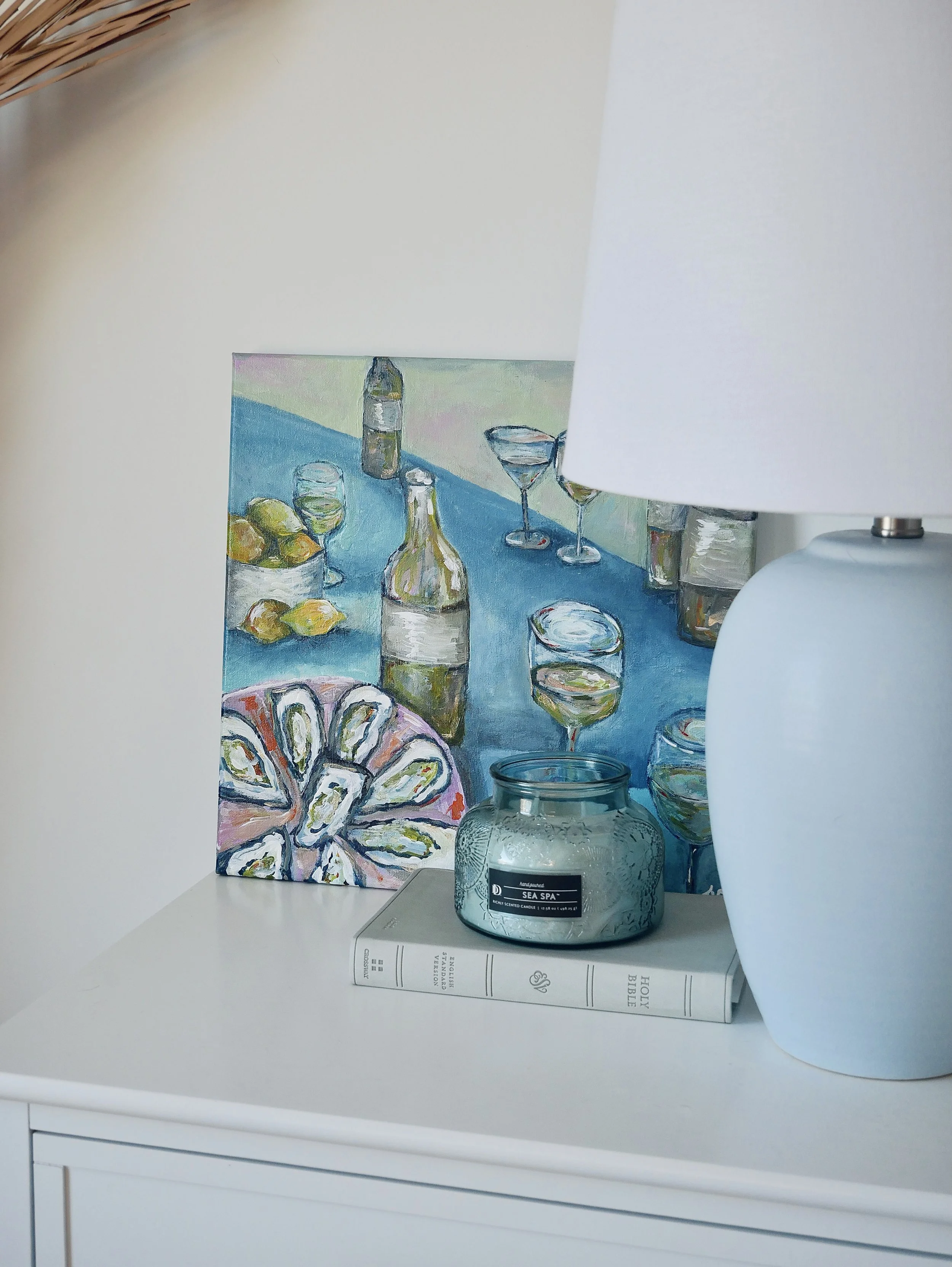 Decorative still life with a colorful painting, a white Bible, a blue candle holder, and a white lamp on a white surface.