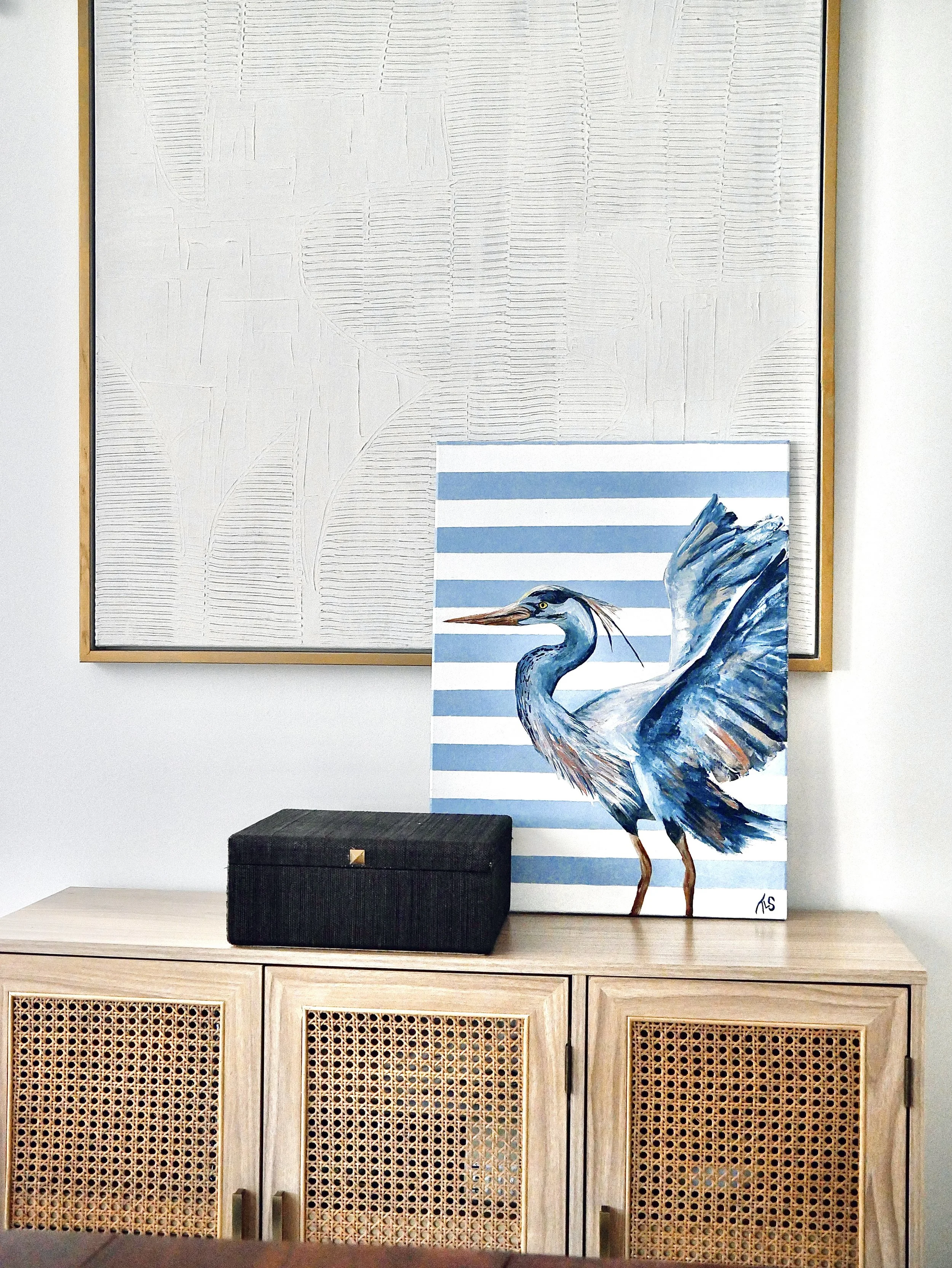 A painting of a blue heron with its wings spread, set against blue and white horizontal stripes, on a wooden cabinet with rattan doors, with a black box on top, and a large textured artwork hanging on the wall behind.
