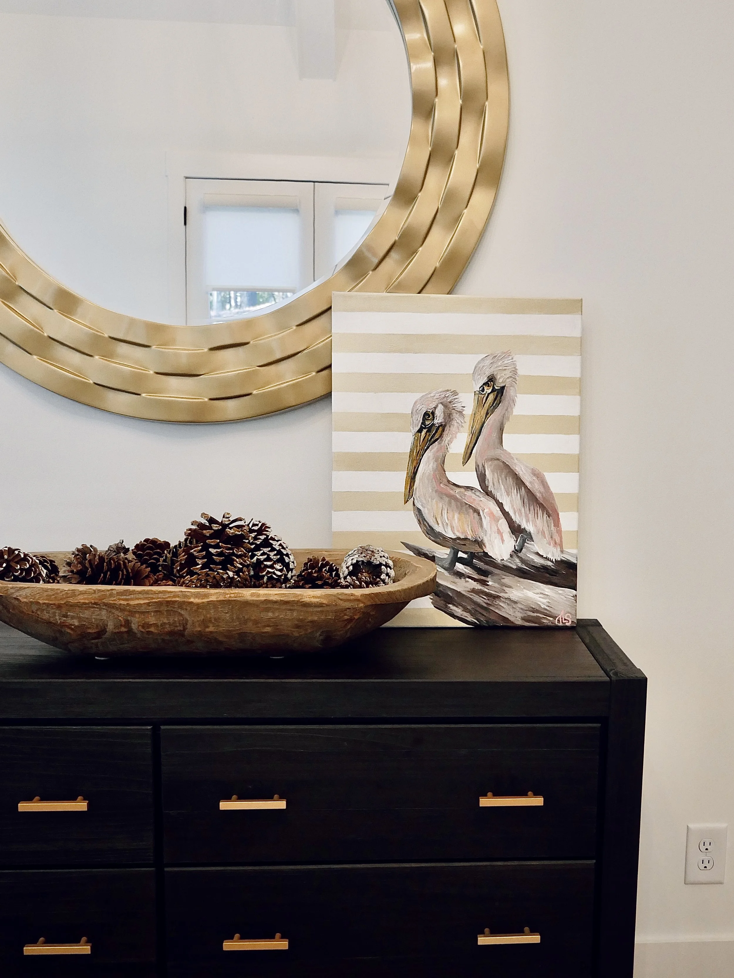 A black dresser with six drawers featuring wooden handles, a decorative wooden bowl with pinecones, a painting of two pink herons with striped beige and white background, and a large round gold mirror on a white wall.