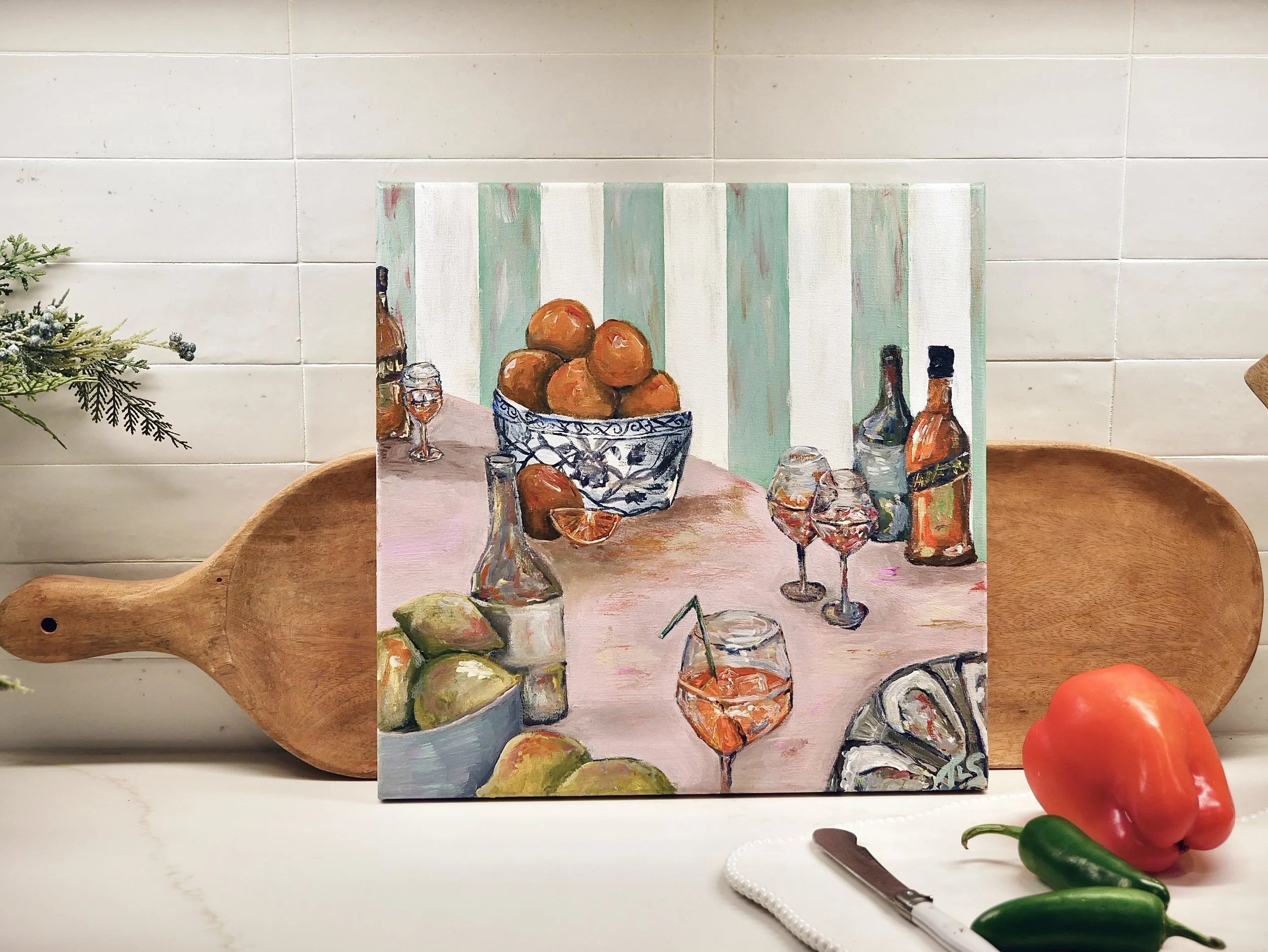 Painting of a table with a bowl of oranges, bottles, glasses of wine, and a lemon on a pink tablecloth, with a striped green and white background.