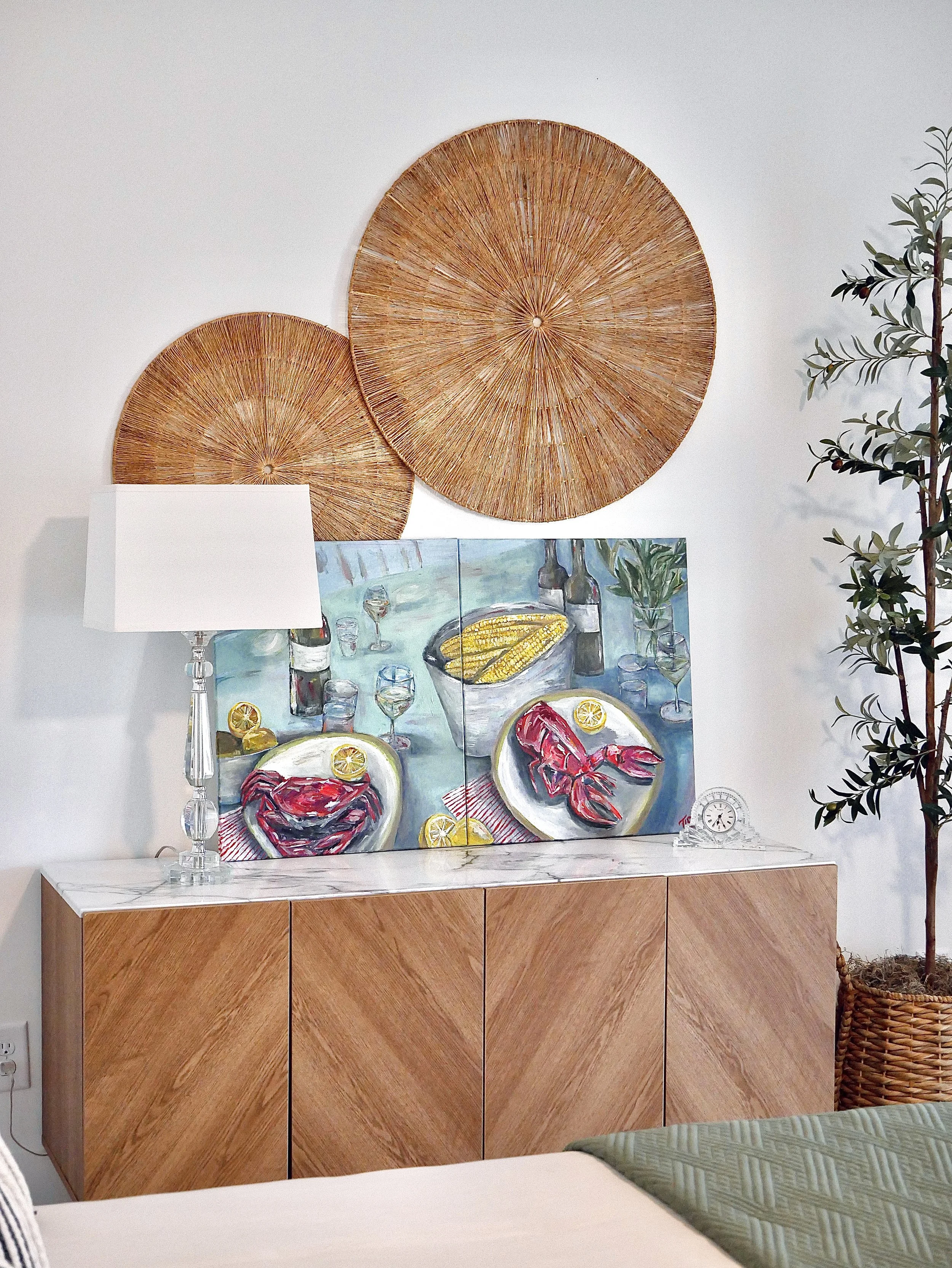 Interior of a room with a wooden cabinet topped with a marble counter, a colorful painting of a seafood meal, a table lamp, a small clock, and decorative wall hangings including woven circular art pieces.