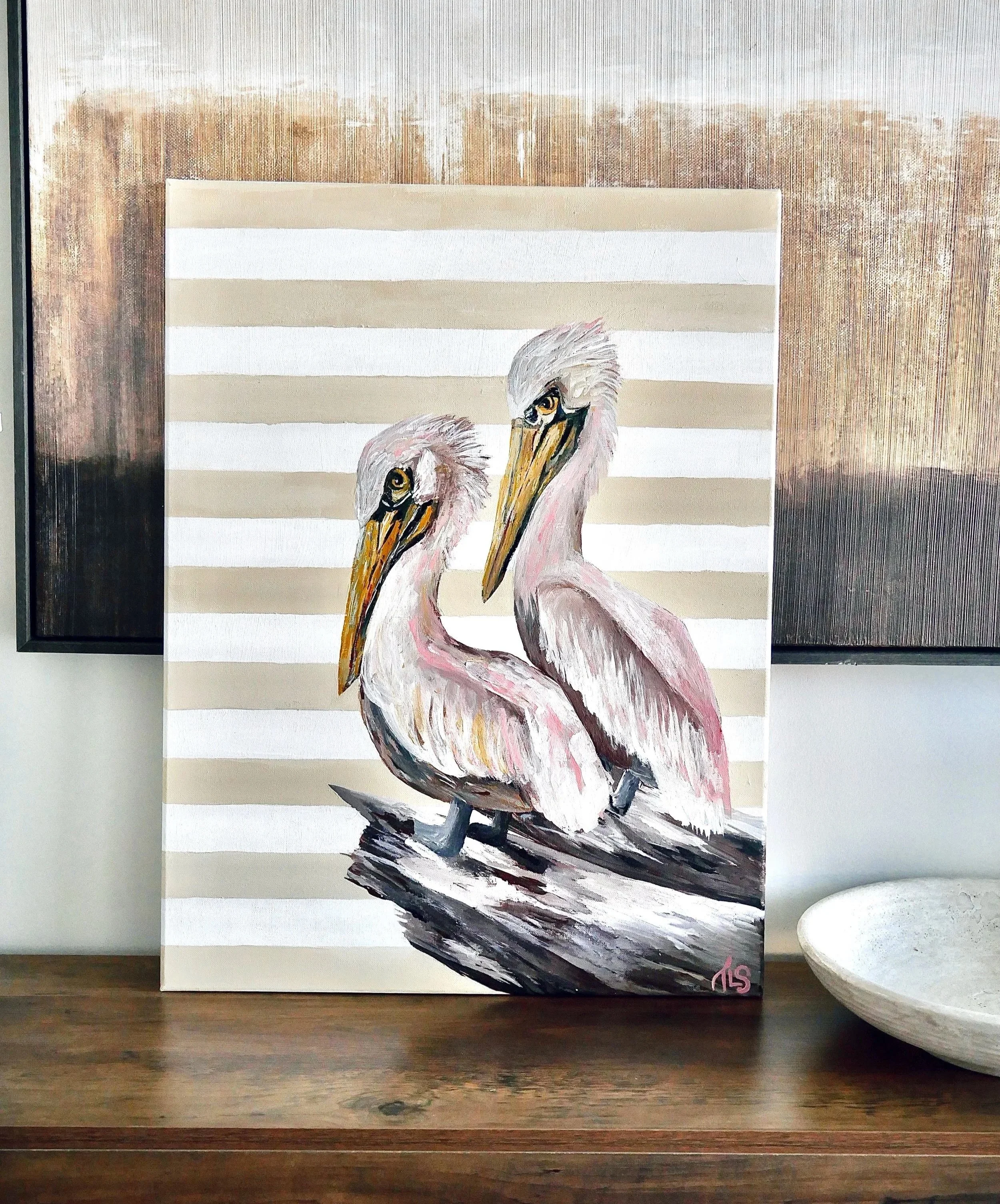 Painting of two pelicans with striped beige and white background, displayed on a wooden surface next to a white bowl.