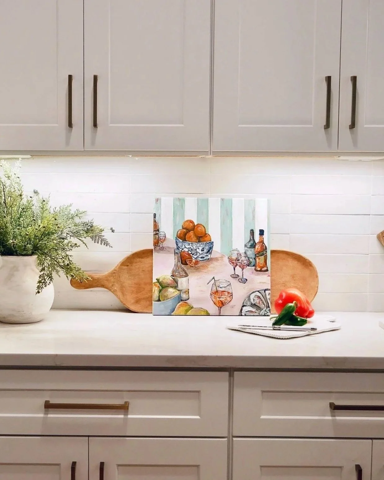 Kitchen countertop with a potted plant, a painting of a still life with oranges and bottles, a red bell pepper, and a cucumber.