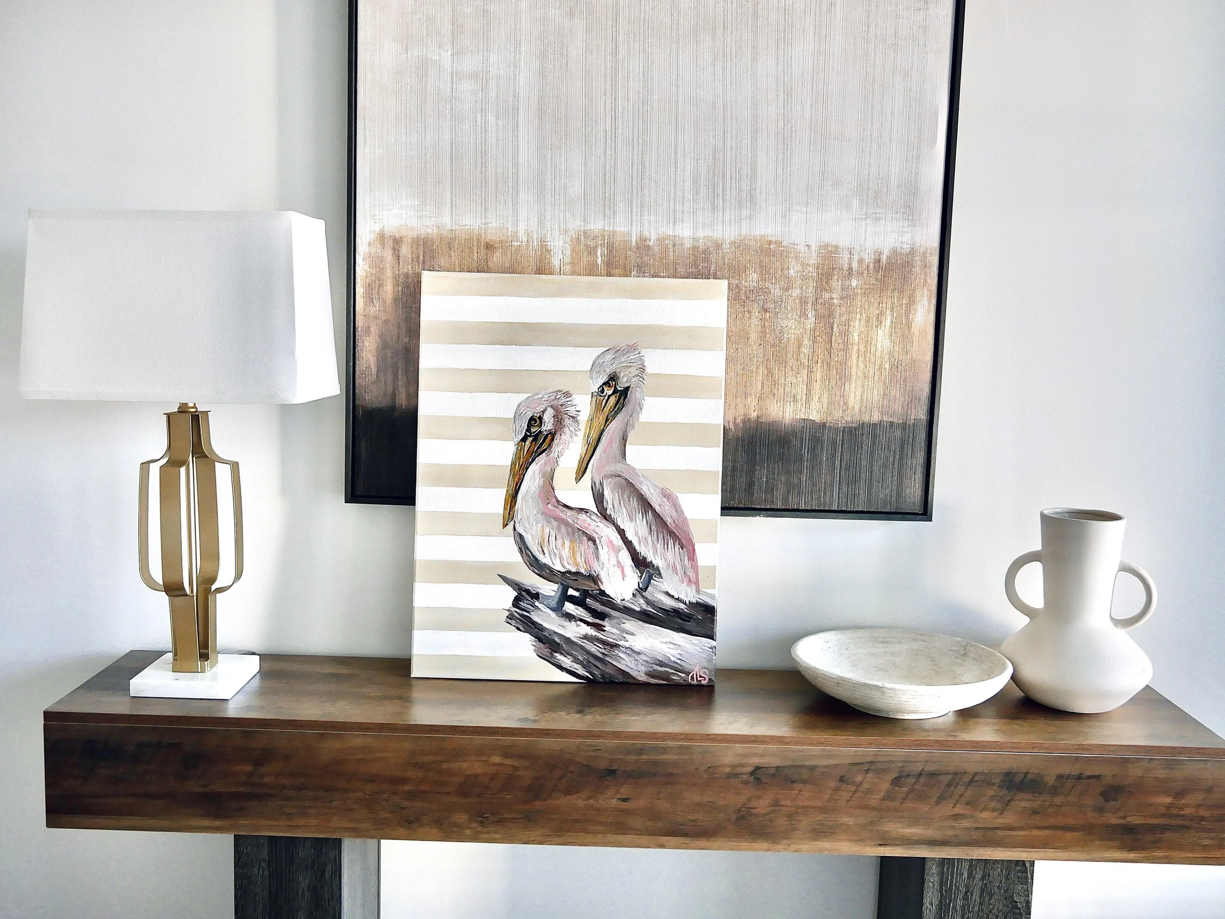 Decorative table with a contemporary lamp, a painting of two pelicans, a bowl, and a vase, against a wall with two abstract art pieces.