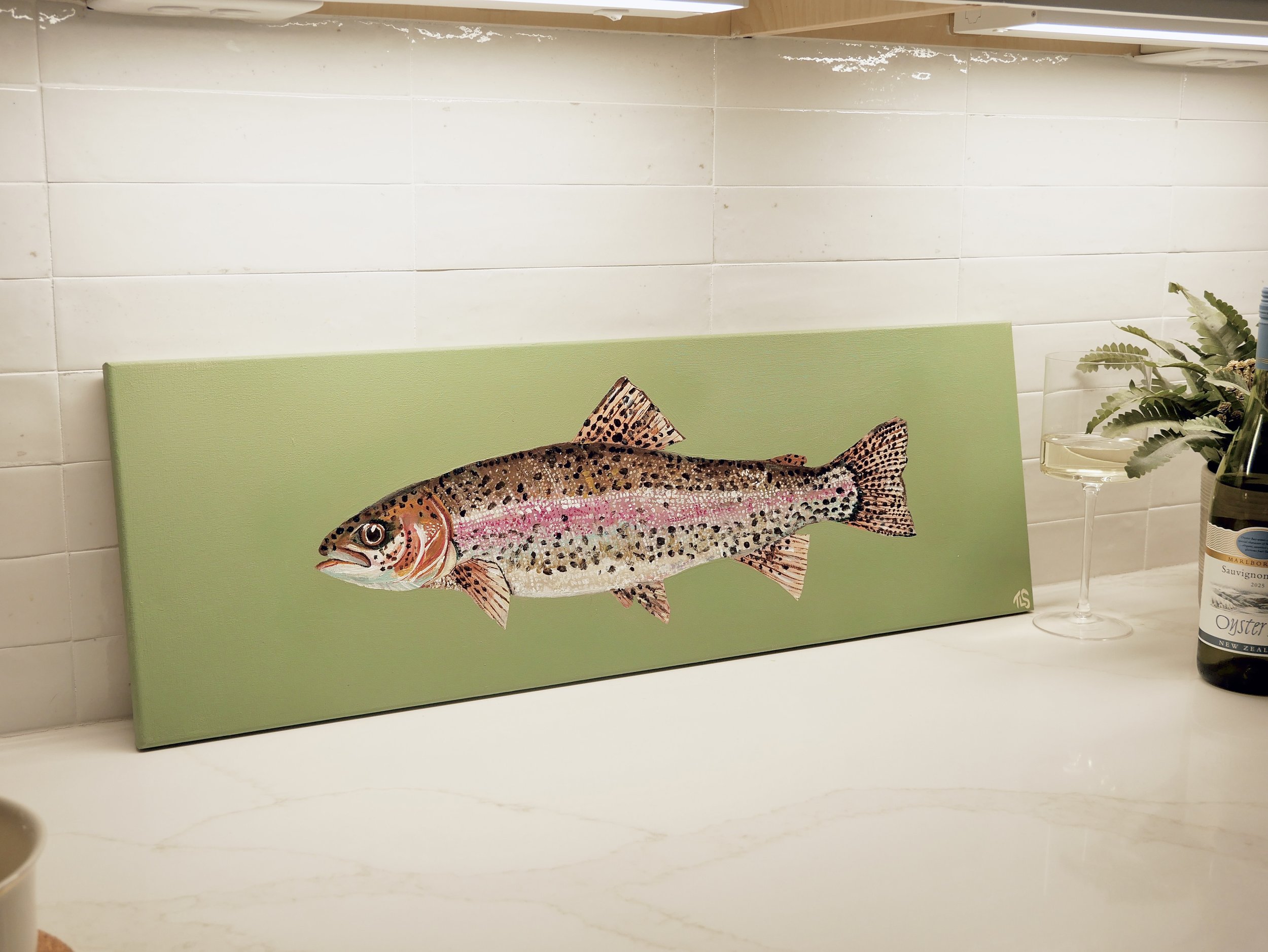 Painted canvas of a rainbow trout on a green background, placed on a countertop with a wine glass and a bottle of wine nearby.