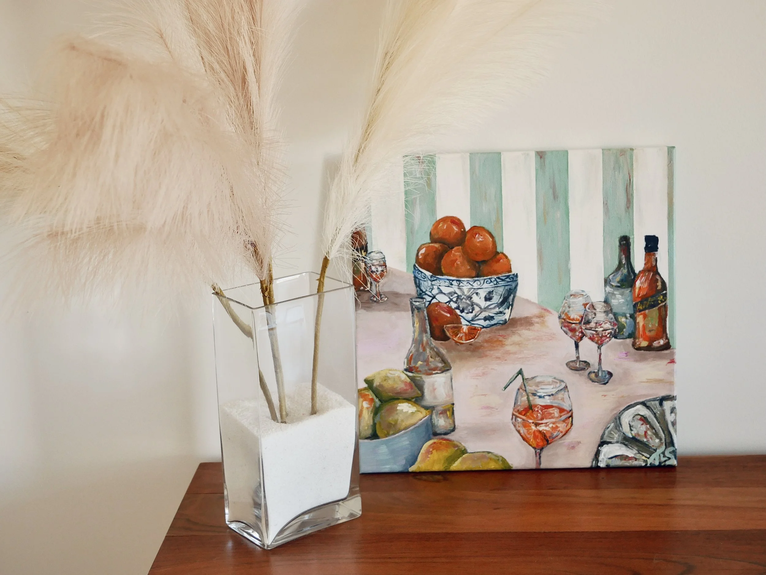 A still life painting of a table with liquor bottles, wine glasses, a bowl of oranges, lemons, and a pitcher, leaning against a white wall with a panel that has vertical stripes in pastel colors.