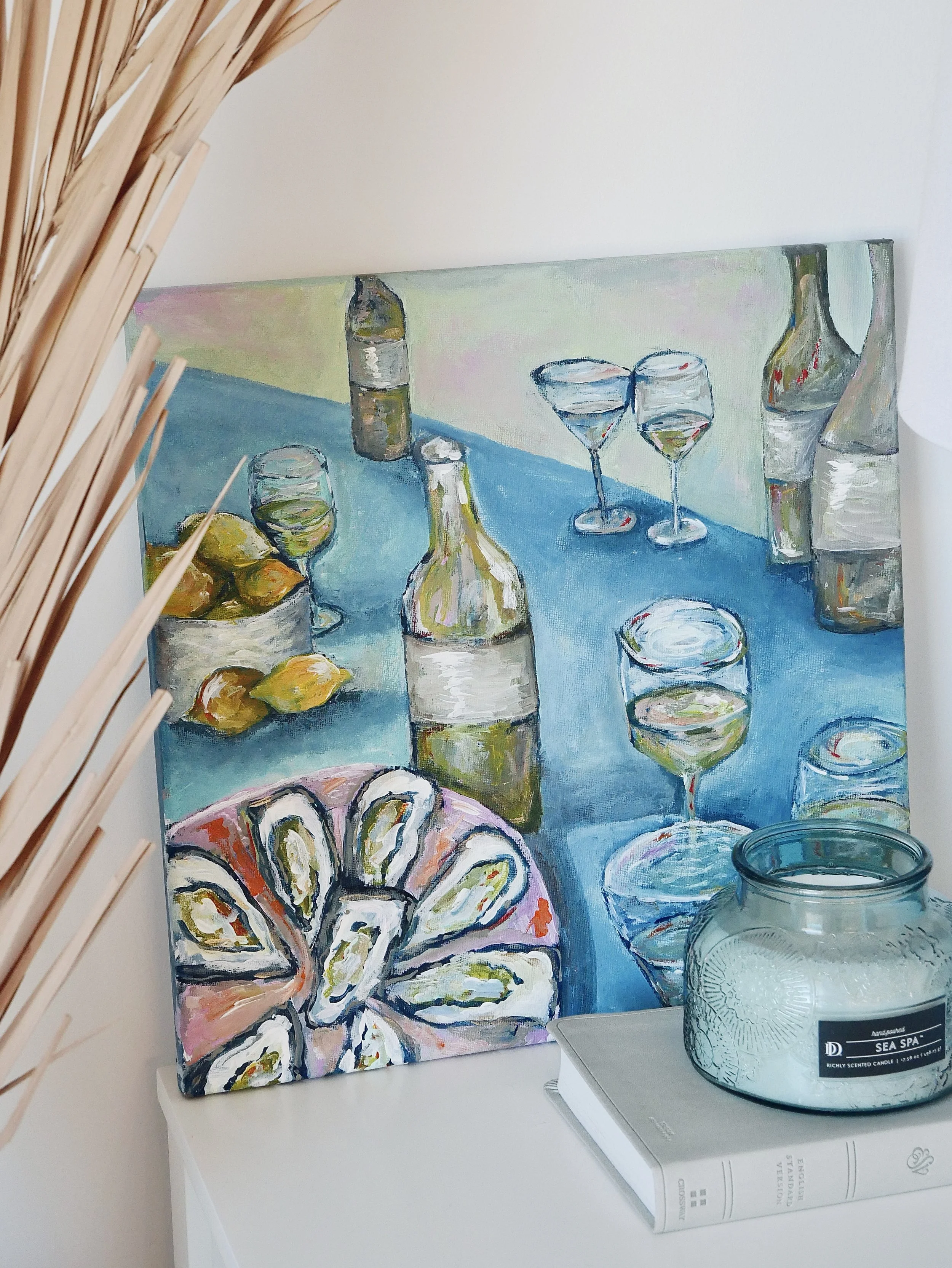 Painting of a table set with bottles of wine, glasses, a bowl of lemons, and a plate of oysters, displayed leaning against a wall with a candle and a book nearby.