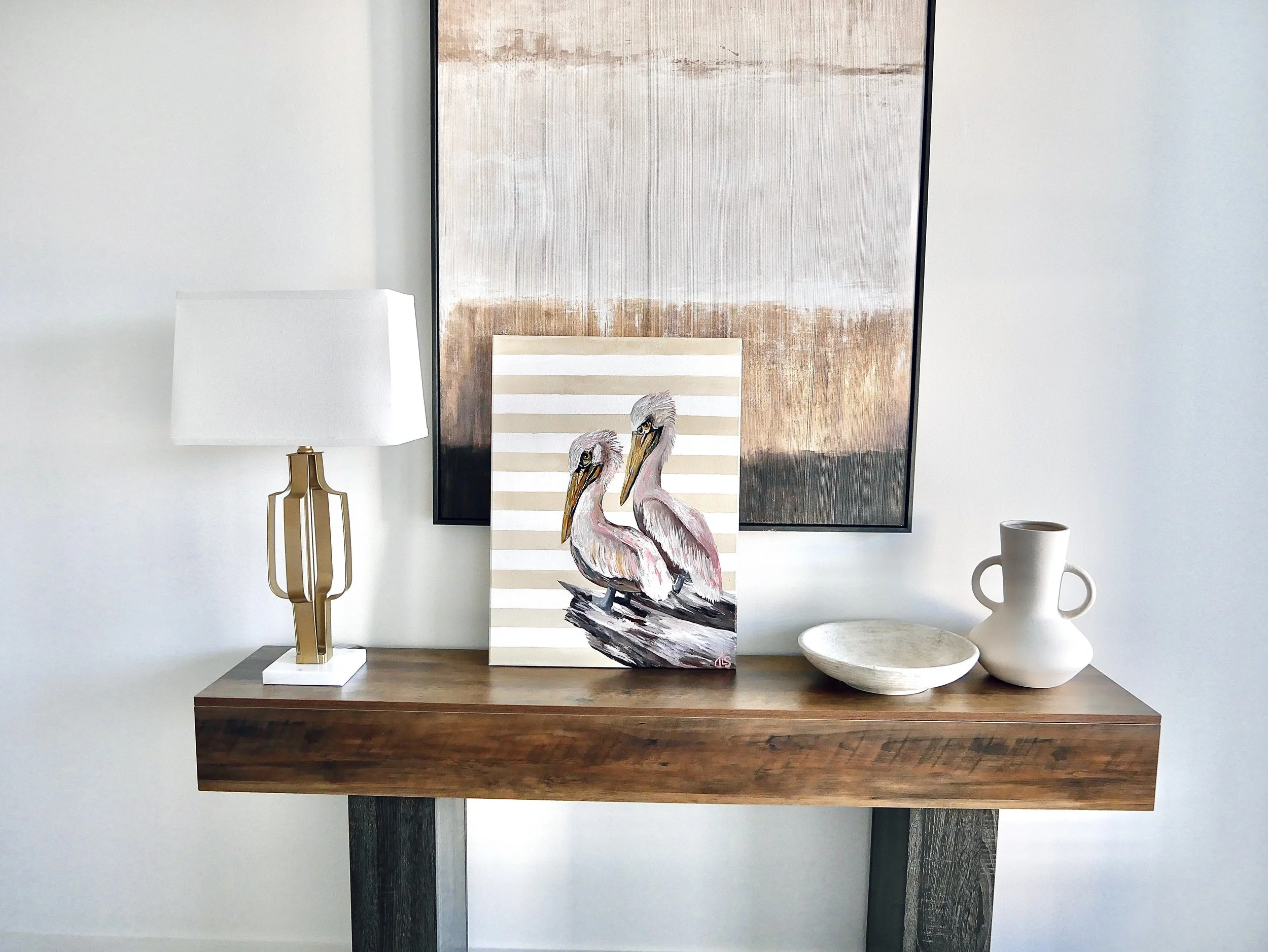 Wooden console table with a modern table lamp, a painting of two flamingos, a white bowl, and a white vase, with an abstract artwork hanging on the wall behind.