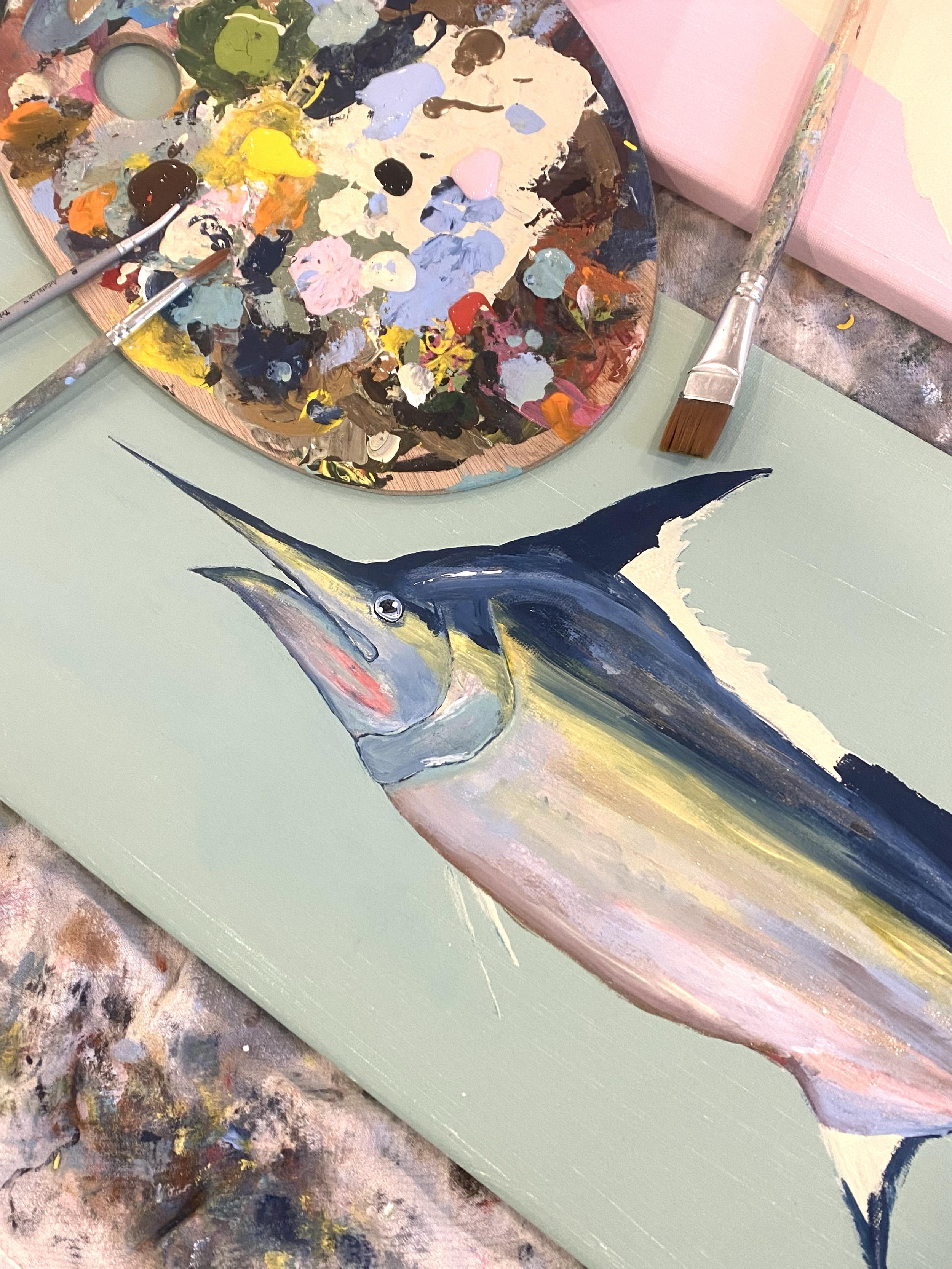 A painting of a marlin fish on a light green canvas, surrounded by painting supplies including a color palette with various paint colors, brushes, and a pink canvas.