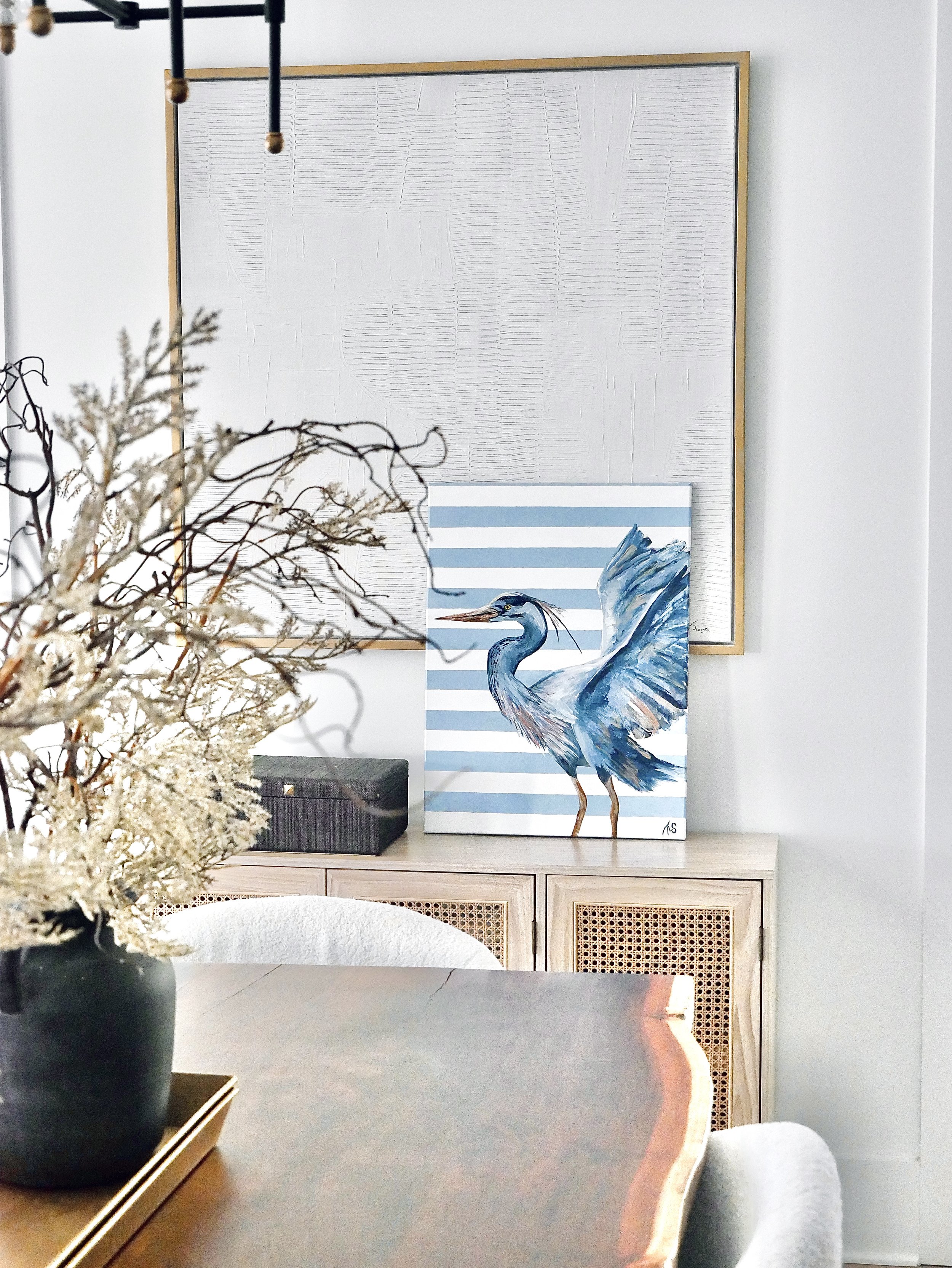 A dining room with a black vase of dried flowers on the wooden table, a white upholstered chair, a cabinet with a woven door, and a painting of a heron with blue and white stripes on the wall.