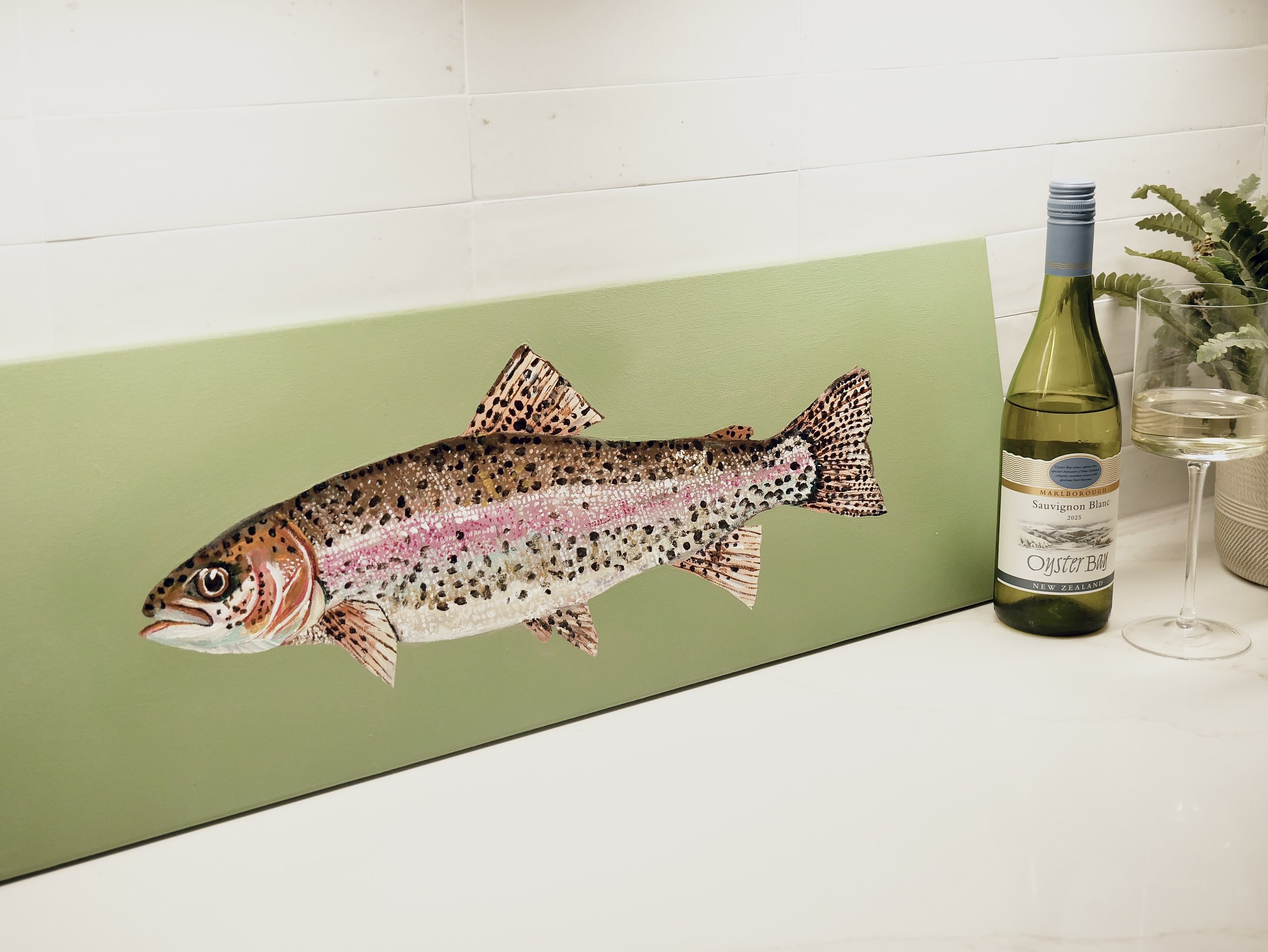 Decorative fish illustration on a green background, with wine bottle, glass of white wine, and plant on a white surface.