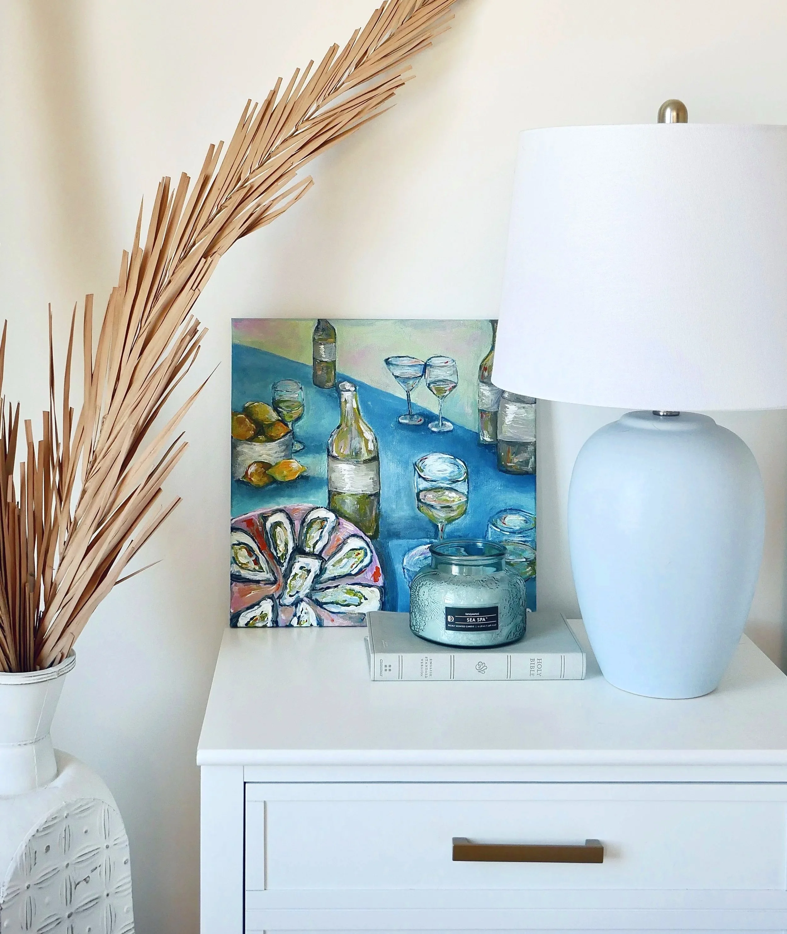 Decorative white lamp on a white dresser with a blue candle, a book, a colorful painting of bottles and wine glasses, and a large white vase with dried brown decorative plants.