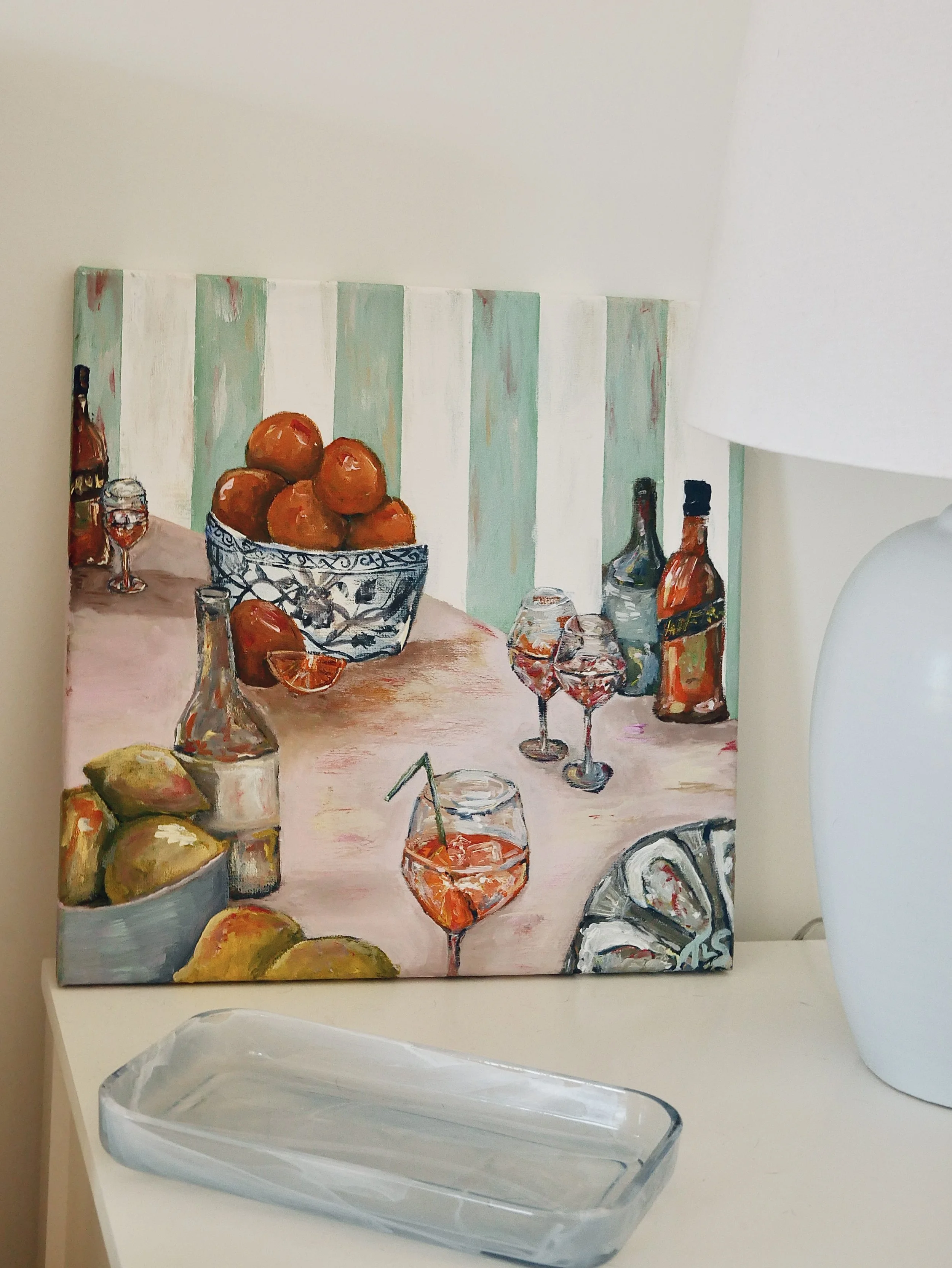 A still-life painting depicting a bowl of oranges, glasses of Aperol spritz with straws, bottles of alcohol, lemons, and a lemon slice on a light-colored table with a striped background.