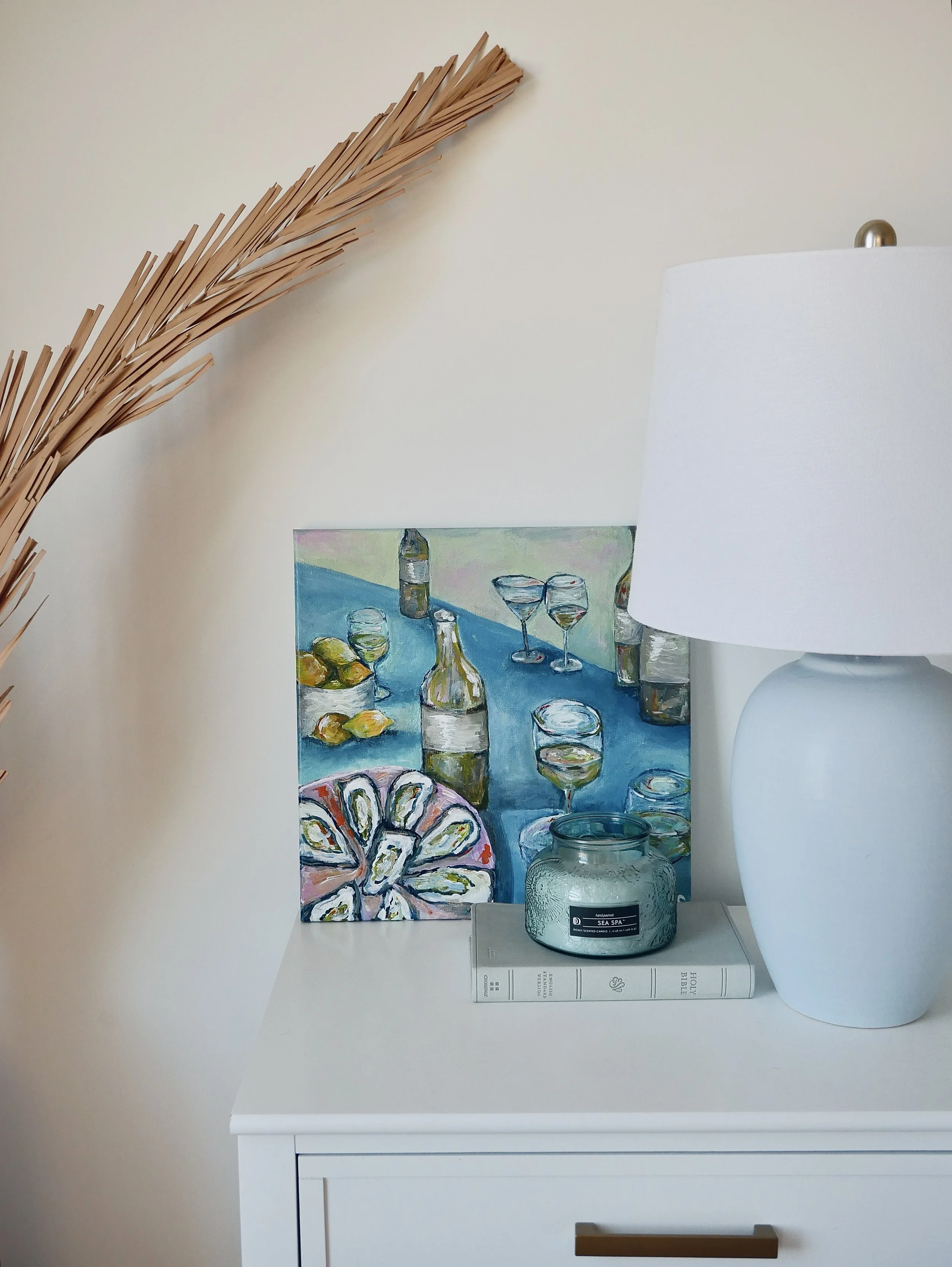 Decorative nightstand with a colorful still life painting, a white table lamp, a green candle in a glass jar, and a white book.
