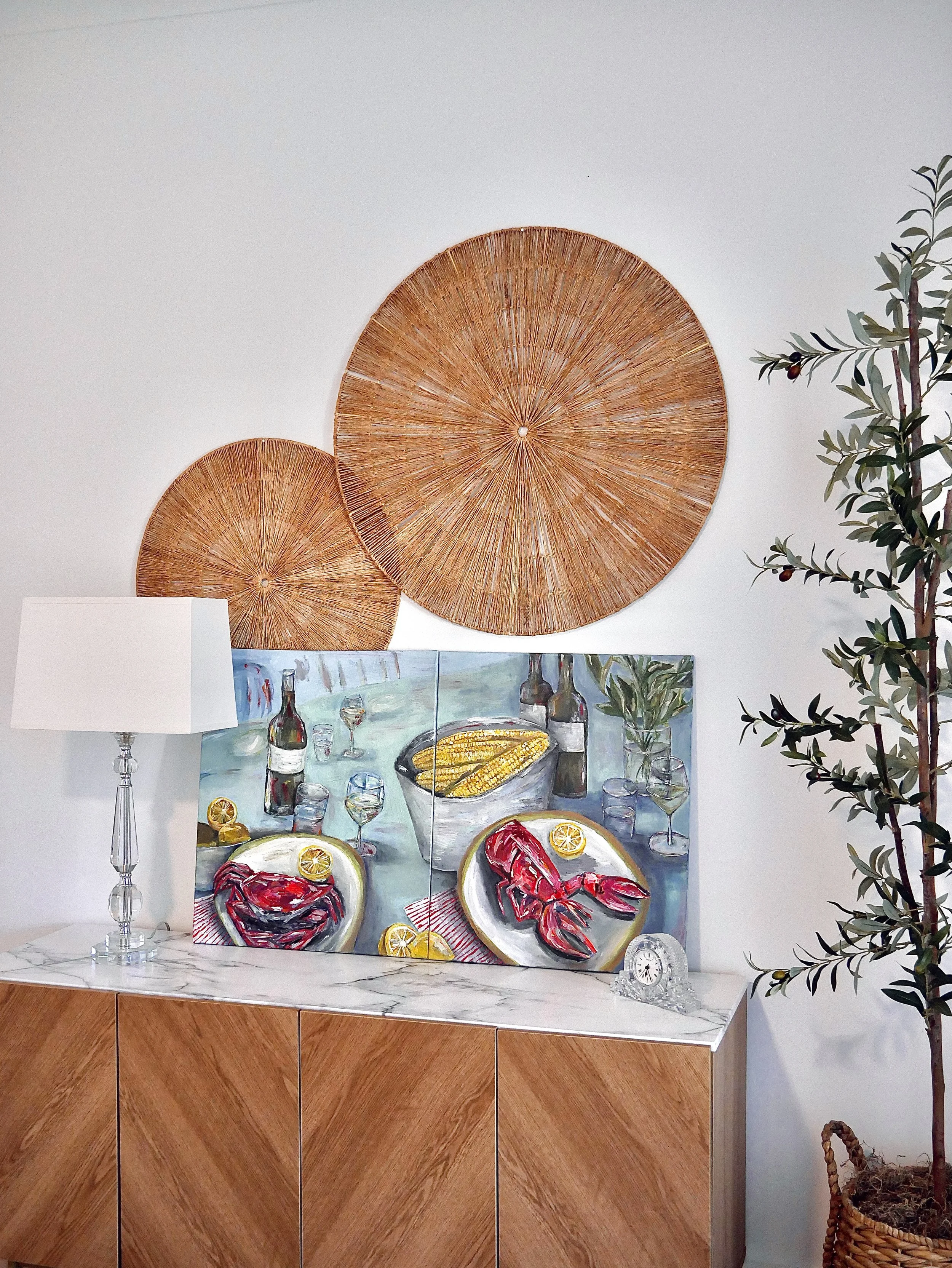 A sideboard with a marble top featuring a painting of a seafood meal with wine bottles and glasses on a table, a white table clock, a white table lamp, and decorative wall art including two large woven circular wall hangings.