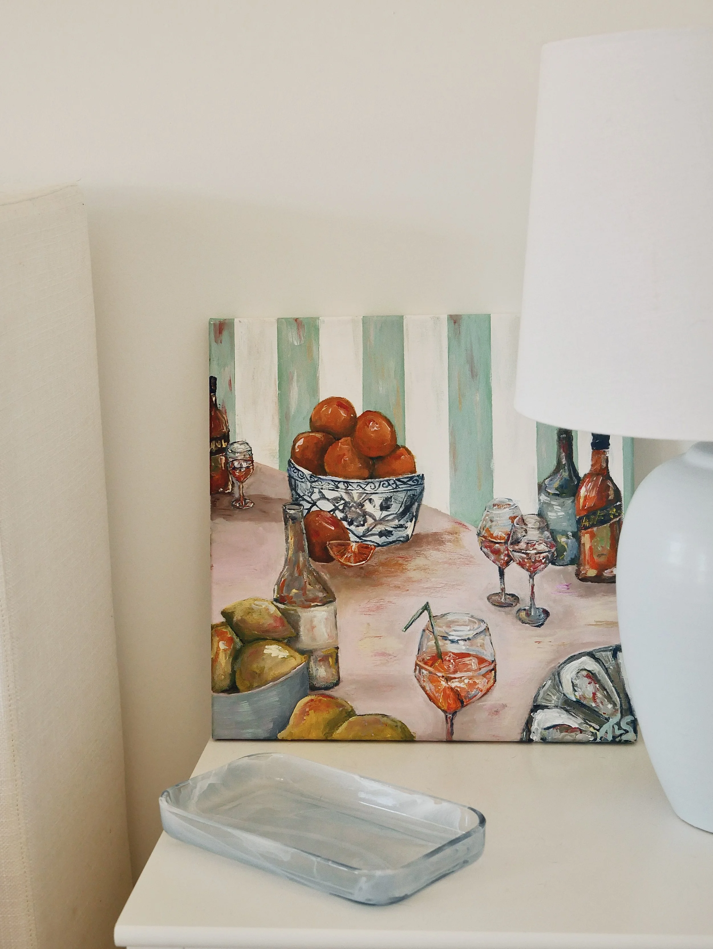 A still life painting of a table with bowls of fruit, bottles, and glasses of wine, leaning against a white wall near a white lamp and a rectangular glass dish.