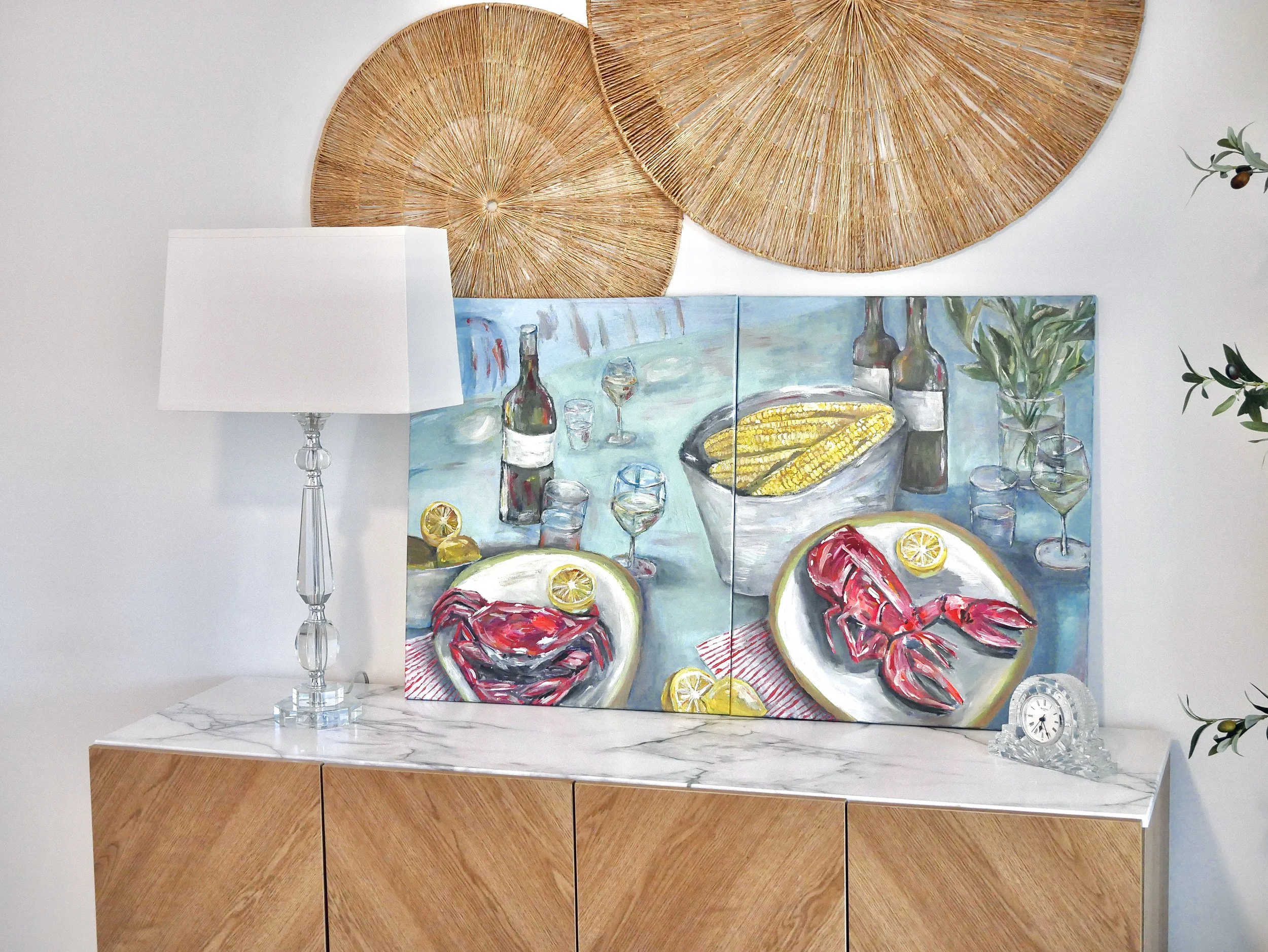 Decorative artwork featuring a still life with seafood, wine bottles, and glasses, positioned on a sideboard with a white marble top. A white table lamp with a clear, sculpted glass base is on the left, and a small clear decorative clock is on the ri