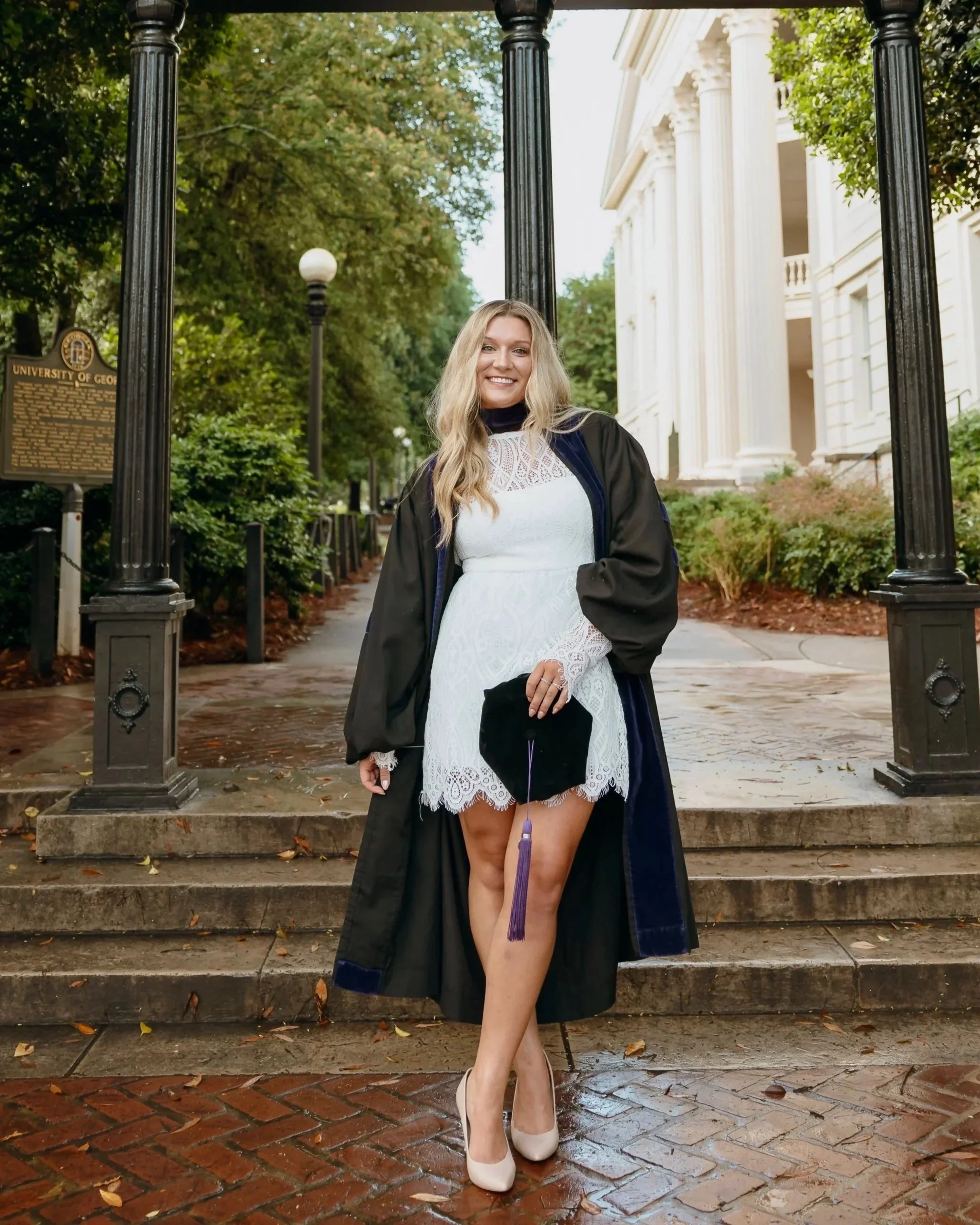 A woman in a white lace dress and high heels holding a graduation cap standing on brick steps in front of a white building with columns and trees.