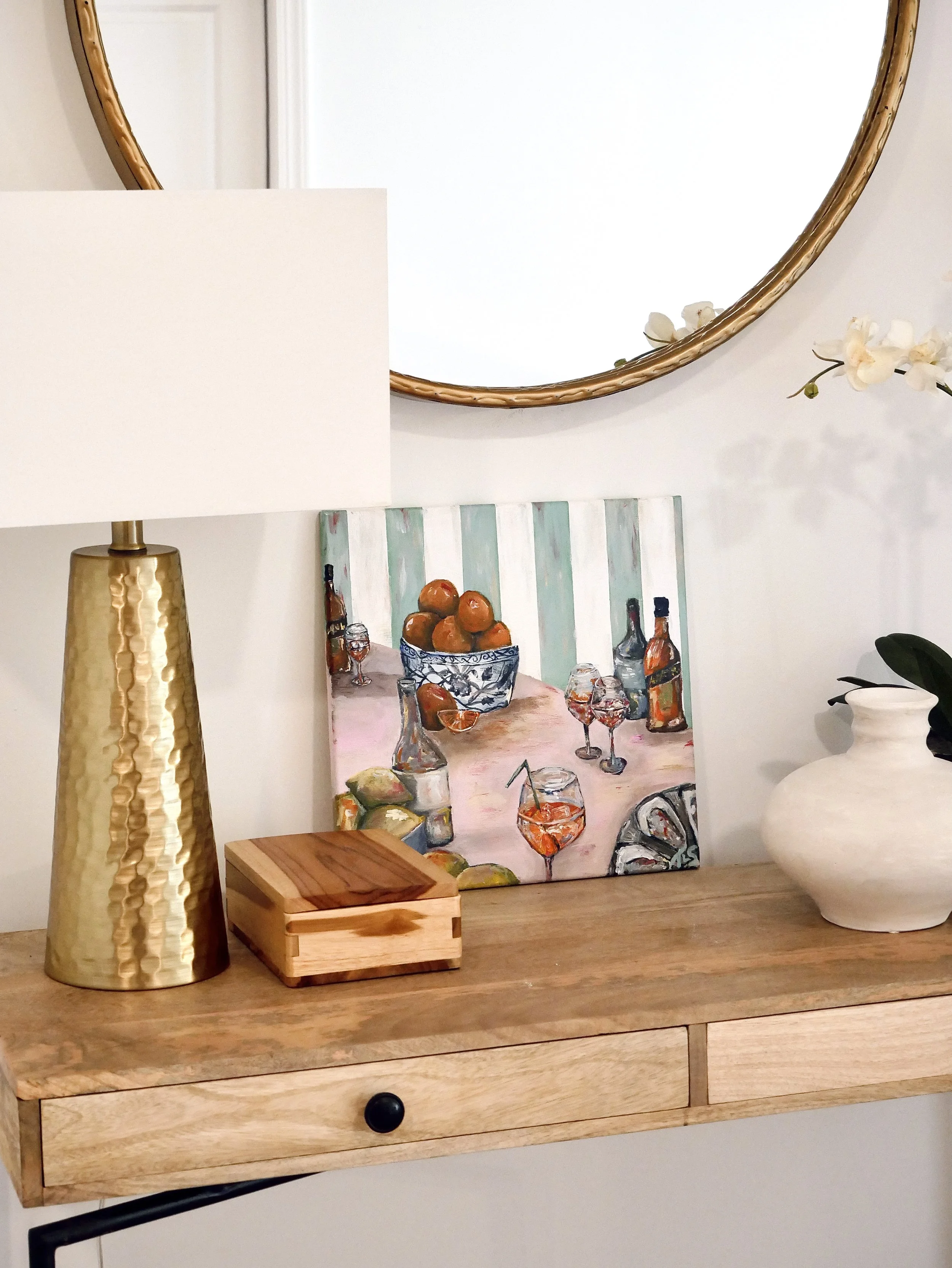 A wooden table with a white ceramic vase, a framed colorful painting of a table with oranges, wine bottles, and glasses, a wooden box with a lid, and a gold textured table lamp with a white lampshade, set against a wall with a round mirror and a whit