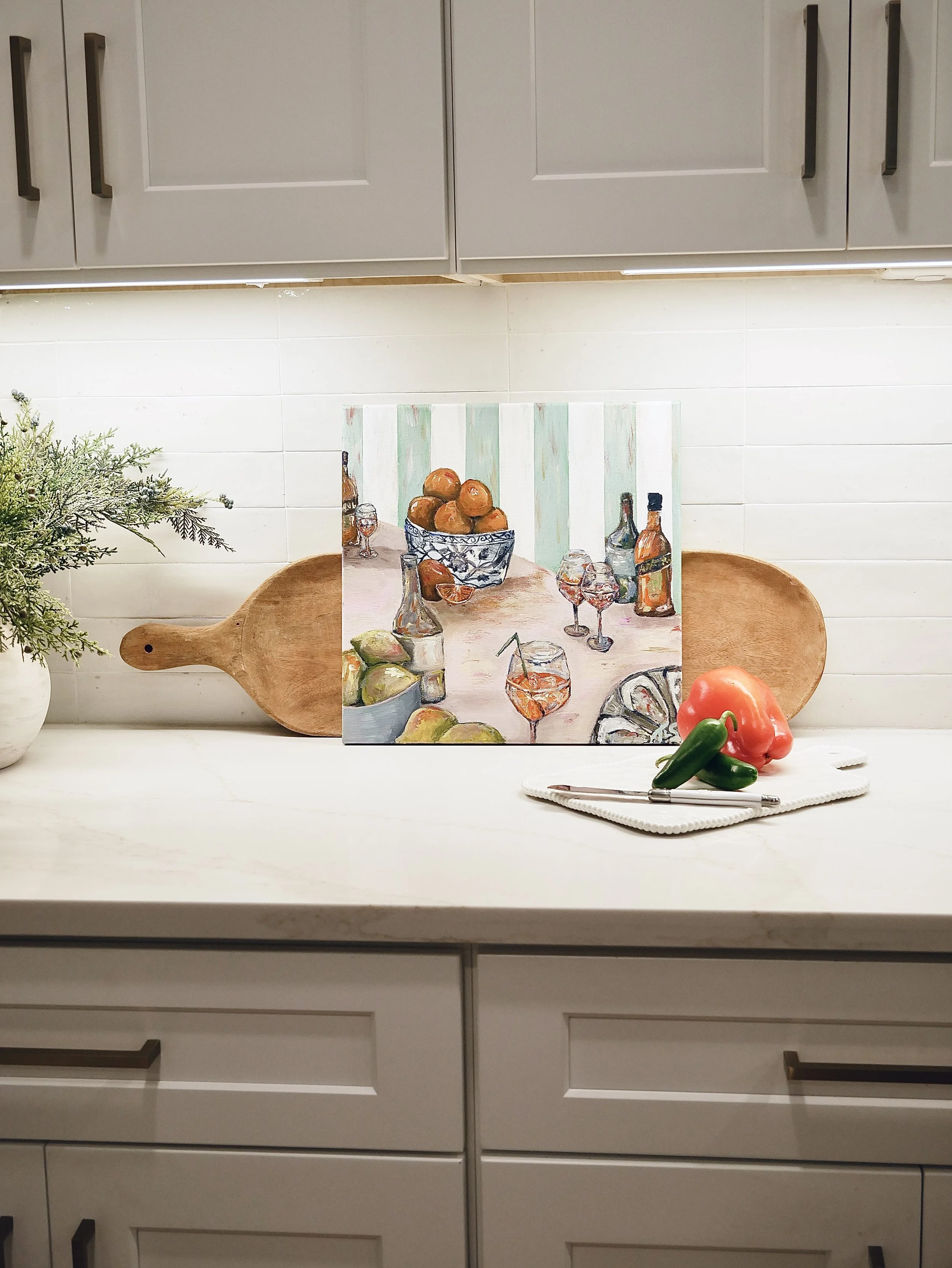Kitchen countertop with a painting of a still life scene, a wooden cutting board, a white plate with a tomato and peppers, and a white notebook with a pen. There are some green foliage on the left side.