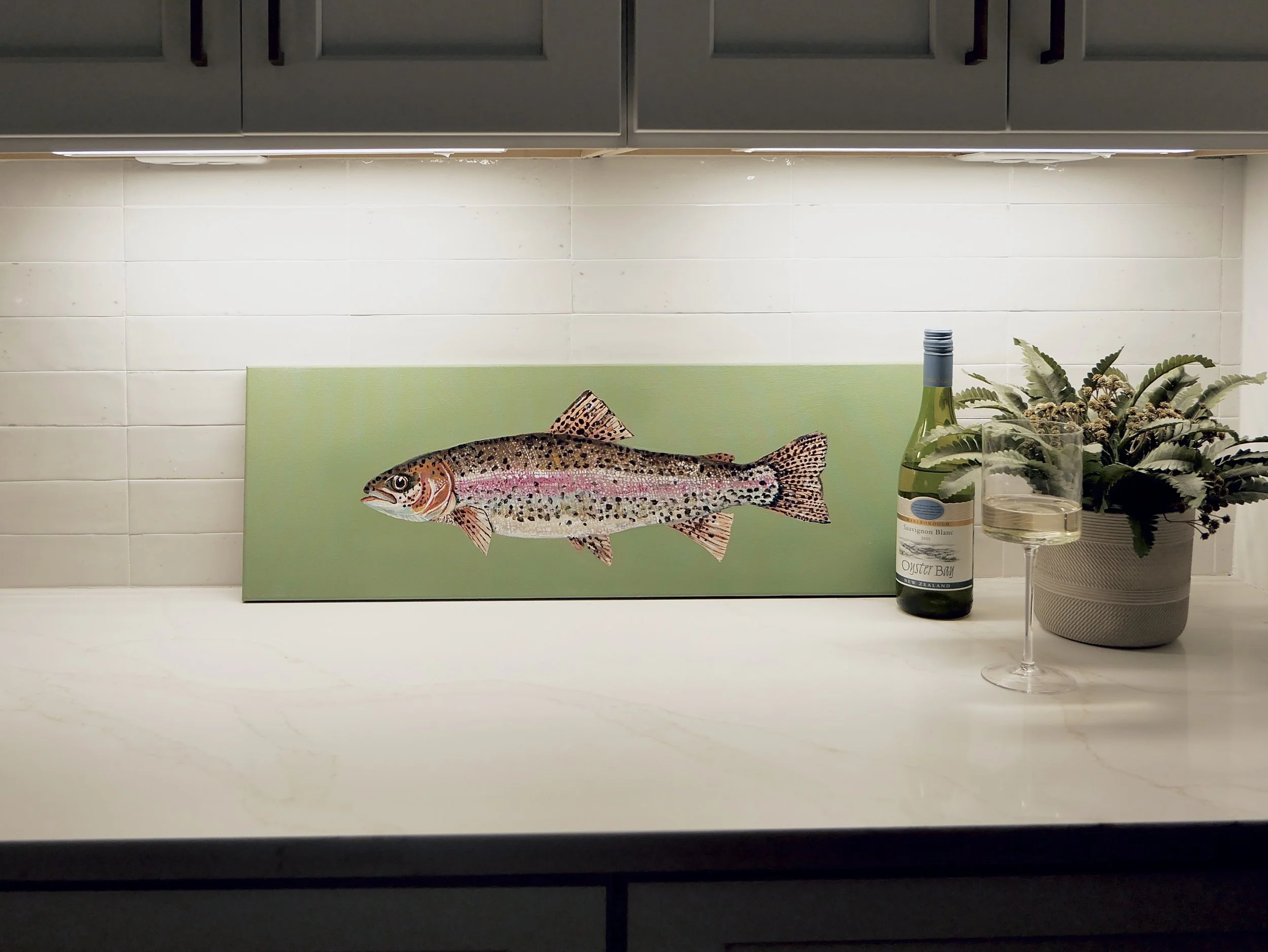 Decorative fish painting on a green background, a wine bottle, a wine glass filled with white wine, and a potted plant on a white countertop in a kitchen setting.