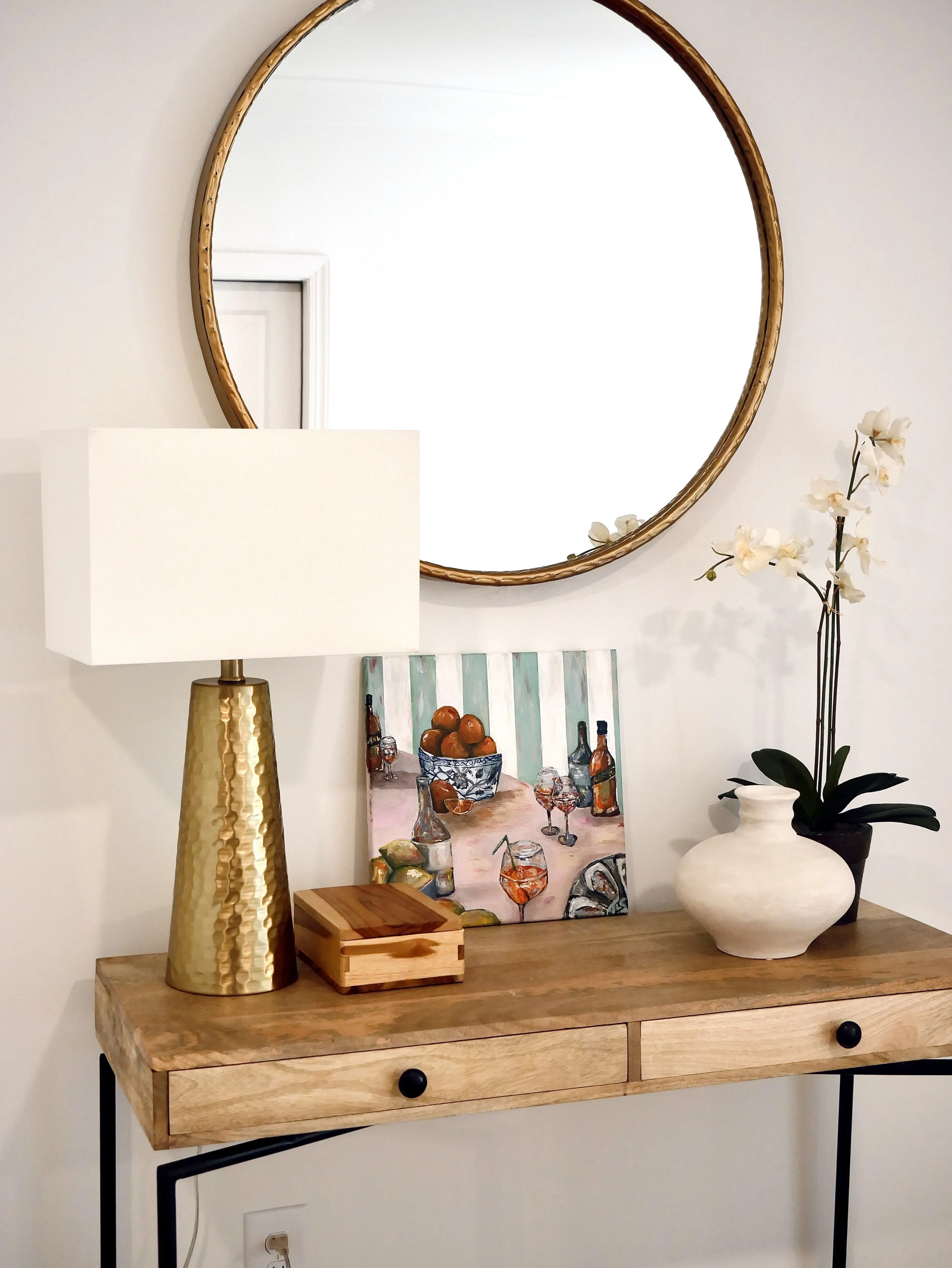 Decorative wooden console table with a gold hammered lamp, a small wooden box, a bowl of oranges, a painting of a dining table with bottles and glasses, a white vase, and an orchid in a pot, with a large round mirror above.