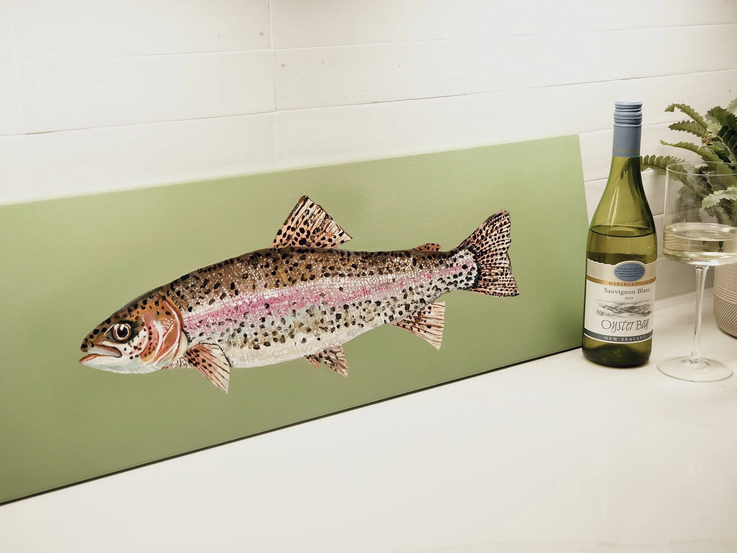 Decorative wall art of a rainbow trout, a bottle of Sauvignon Blanc wine, a wine glass, and a potted plant on a white countertop.