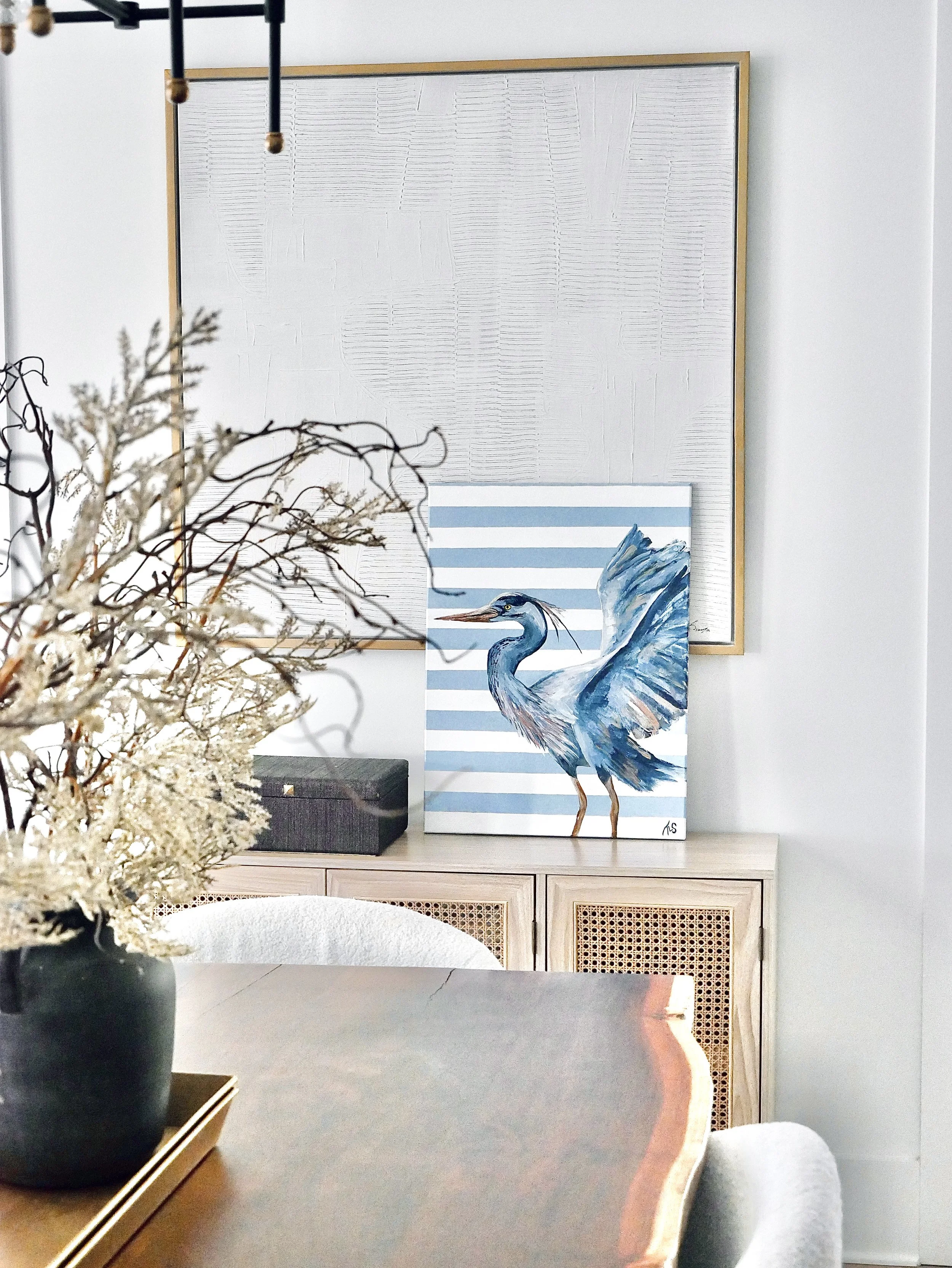 A close-up of a table with a black vase holding dried flowers in the foreground, a wooden cabinet with cane doors and a decorative artwork of a heron, and an empty textured white wall with a gold frame in the background.