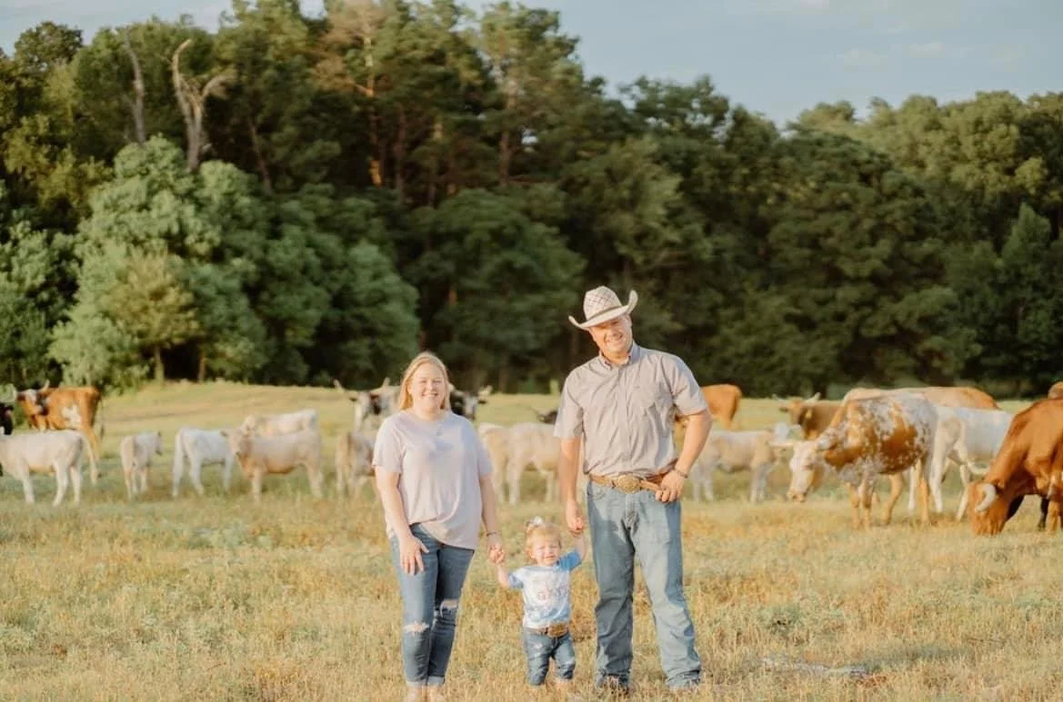 A family of three standing on a farm with cows grazing in the background and trees behind them, enjoying a sunny day.