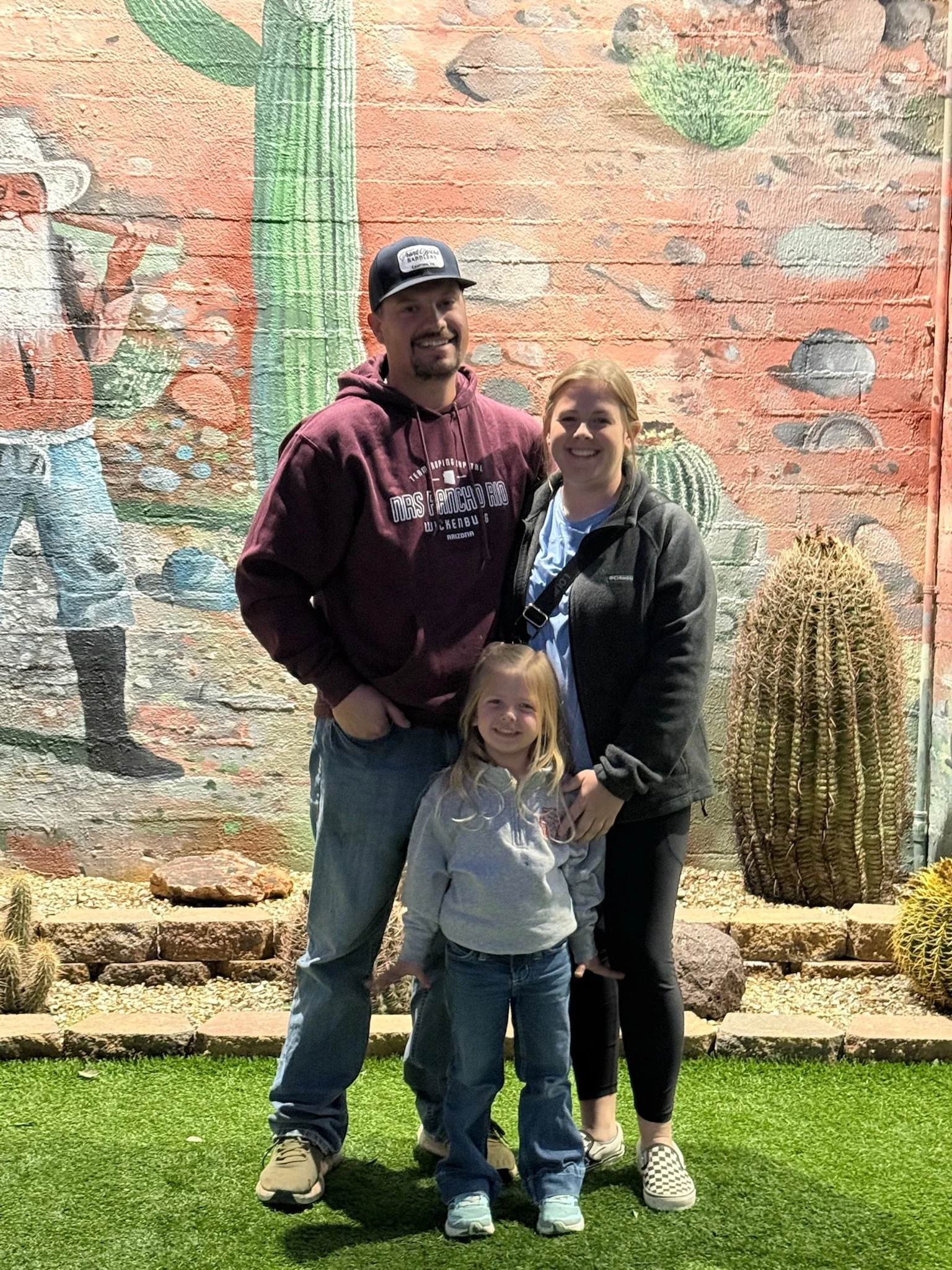 A family of three standing outdoors in front of a colorful desert-themed mural with cacti and rocks, smiling at the camera.