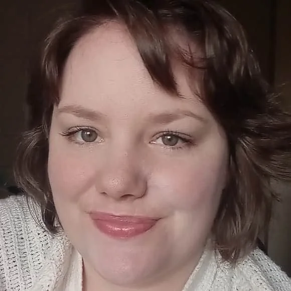 Close-up of a woman with short, wavy brown hair and light-colored eyes, smiling gently at the camera.
