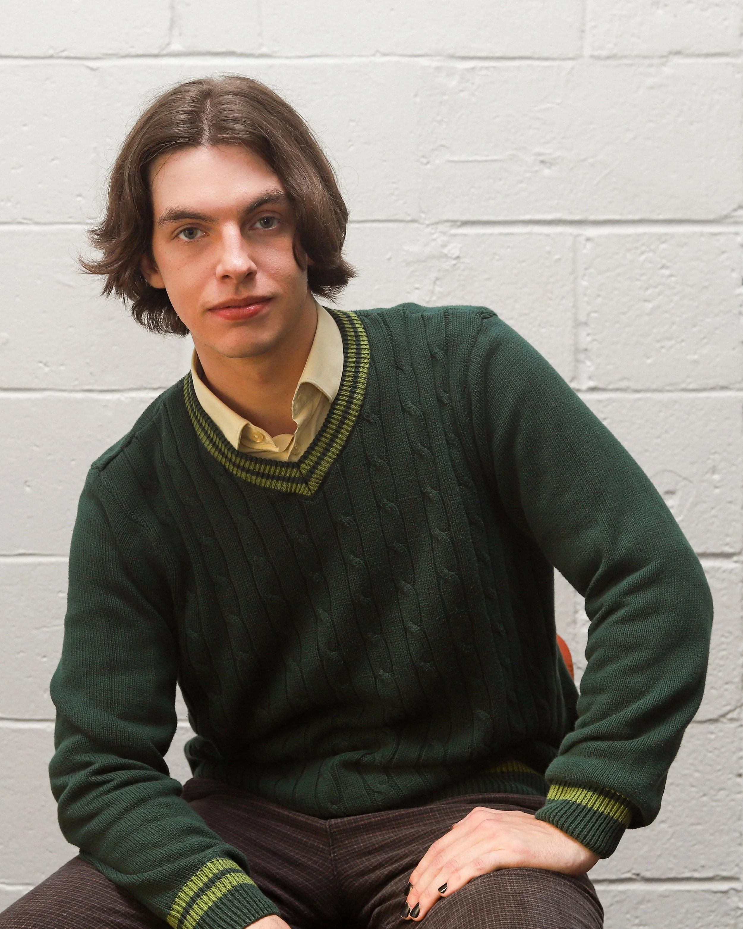 Ryan, a non binary performer, faces the camera while leaning their body to the left of frame. They have chin length brown hair, and is wearing a green sweater, yellow button down, and brown pants. The background is white brick.