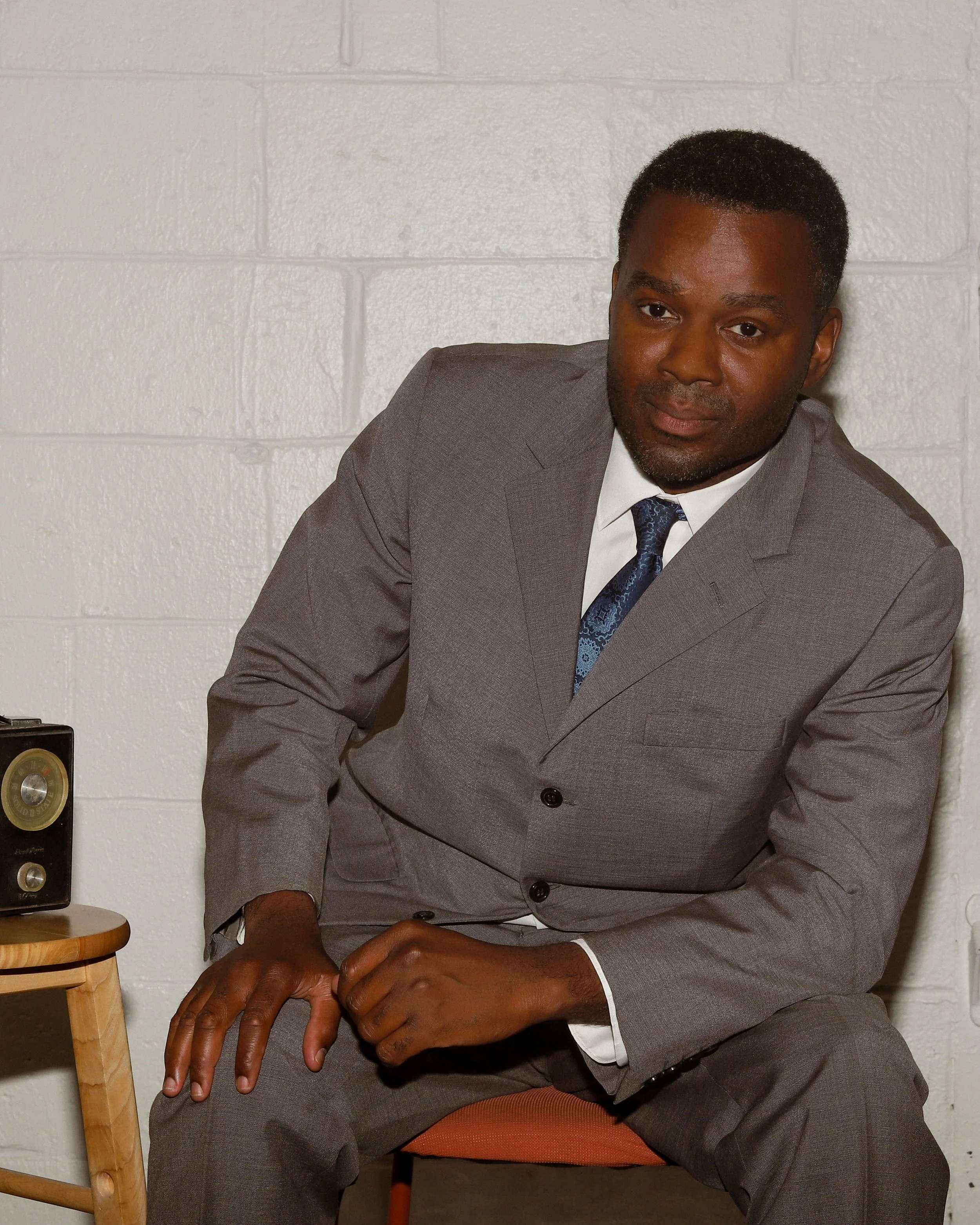 Jason, a black man, sits on an orange chair and leans his left arm against his leg, tilting his body. He wears a grey suit with a blue tie. The background is white brick.