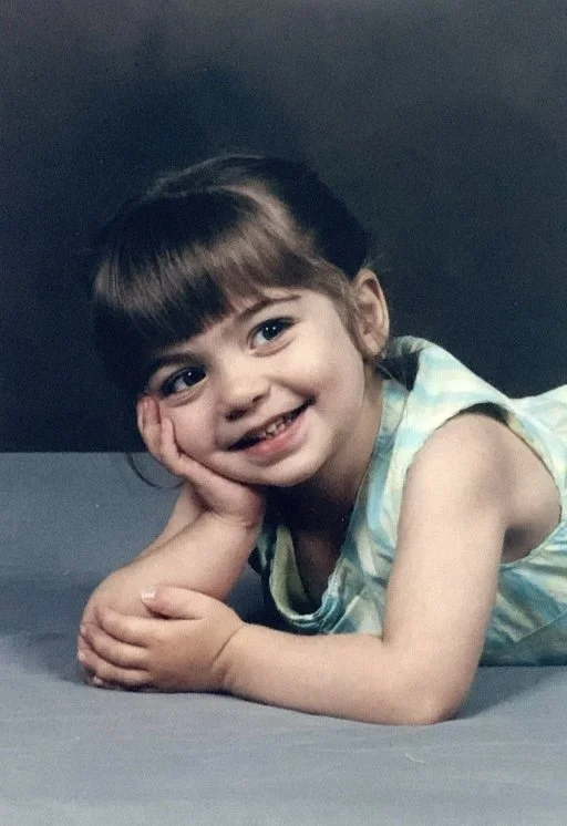 Little Jaelynne is smiling at the camera. She is posing with her cheek on her hand, and her other hand resting on her elbow. She is wearing a green dress.