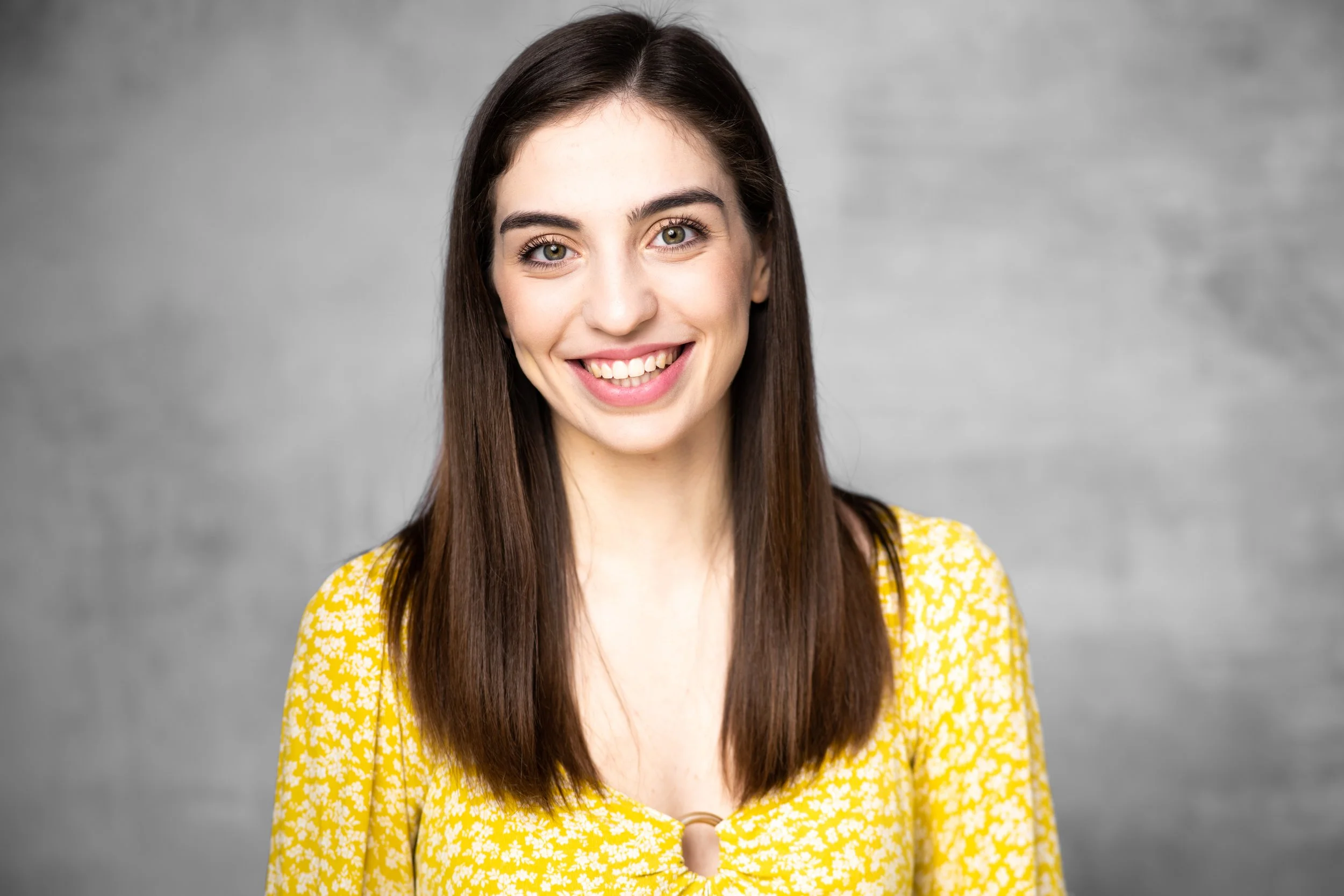 Jaelynne, a white woman, is smiling at the camera. She has long brown hair, and is wearing a yellow blouse. The background is light grey.