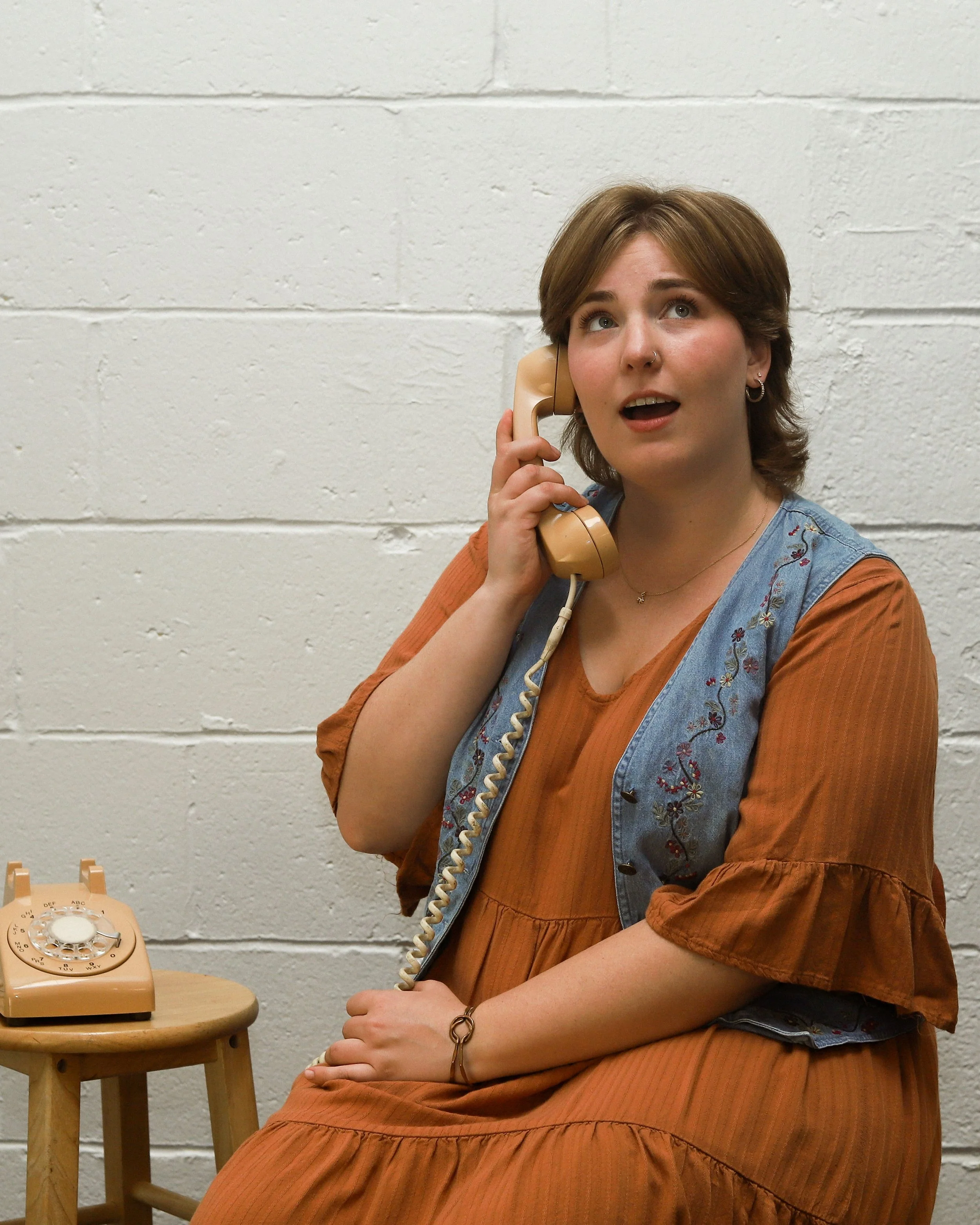 Annie, a white person, sits and holds a rotary phone to her ear. They have an engaged, happy expression on their face. She is wearing an orange dress and denim vest. The background is white brick.