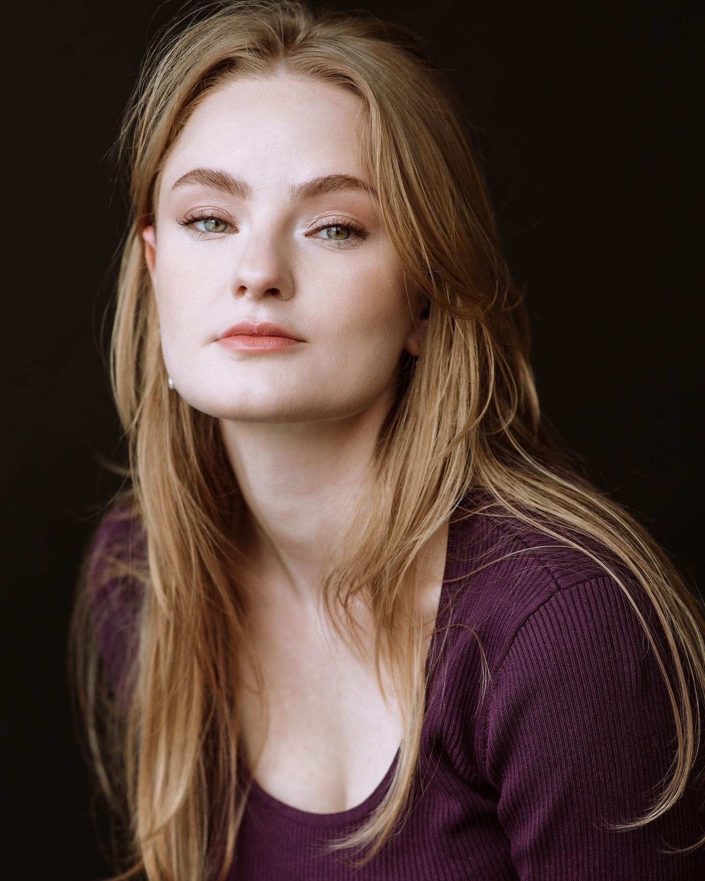 Katelyn is a white woman with long blonde hair, and green eyes. She is leaning towards the camera, wearing a burgundy shirt. She is in front of a black background.