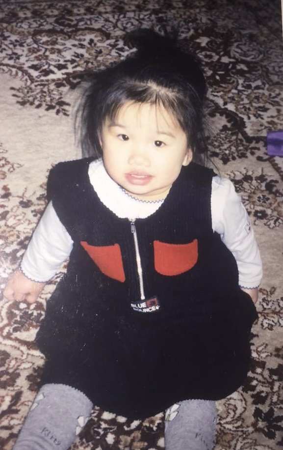 Little Joanne is smiling at the camera, wearing a black overalls dress and a white long sleeve shirt.