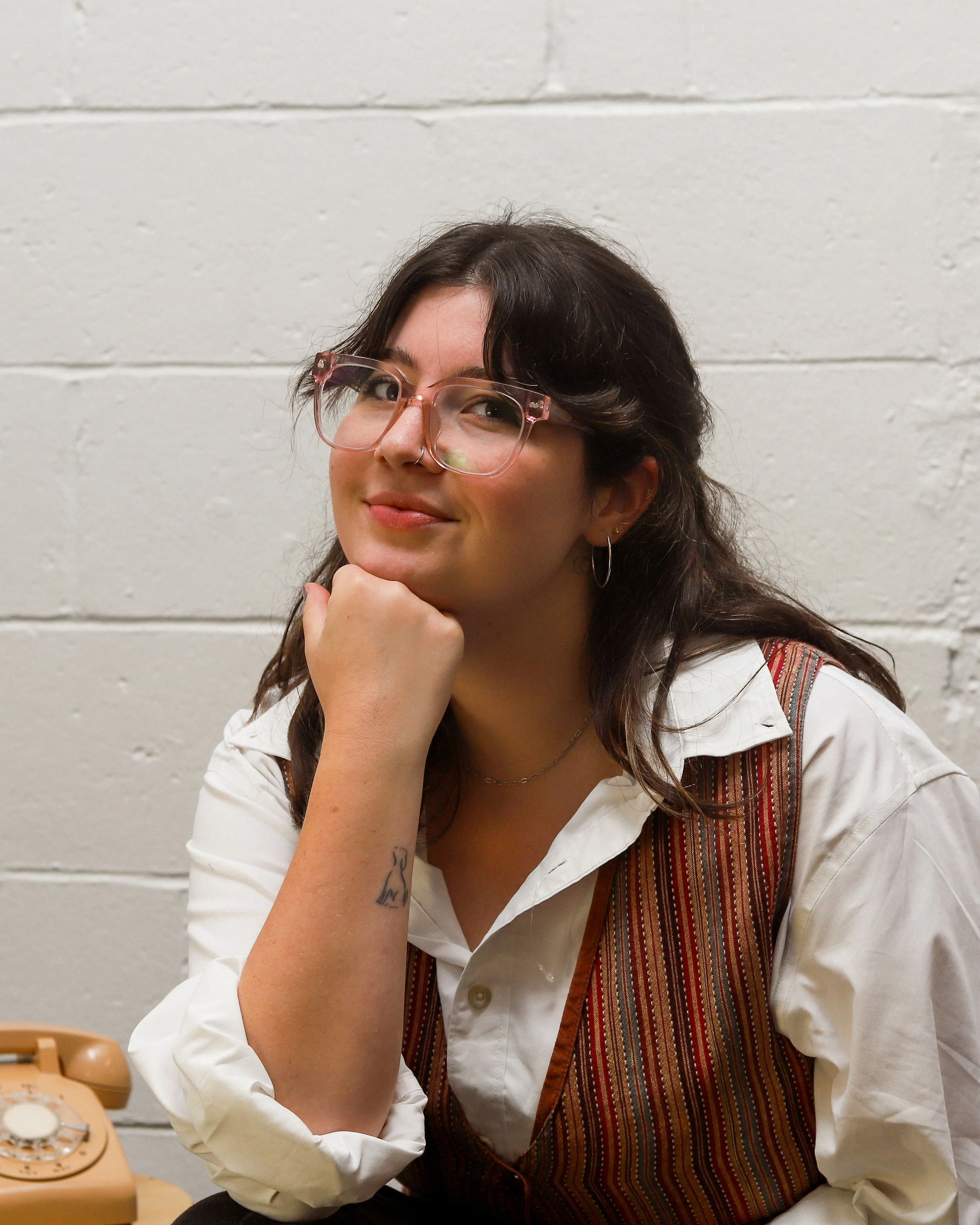 Alexa, a white woman, smirks at the camera, her chin resting on her fist. She has shoulder-length brown hair, glasses, and a nose ring. She is wearing a white button down and a multi-coloured vest. The background is white brick.